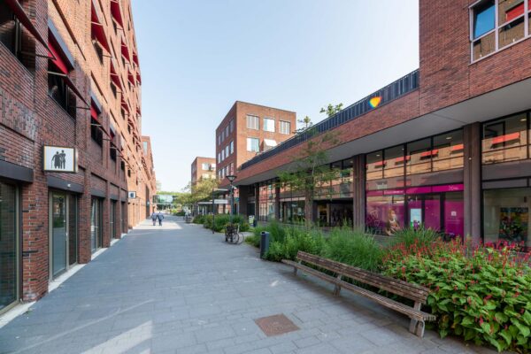 Moderne straat aan de Maasstadweg met rode bakstenen gebouwen, winkels, groenstroken en bankjes.
