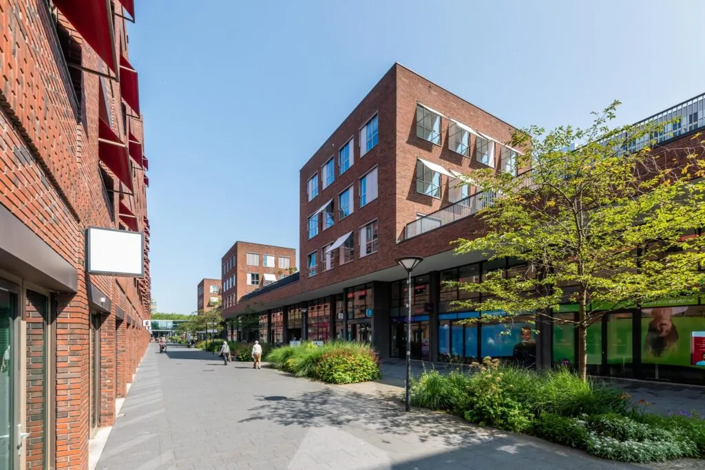 Moderne bakstenen gebouwen aan de Maasstadweg met winkels op de begane grond en een groenstrook langs een voetgangersgebied.
