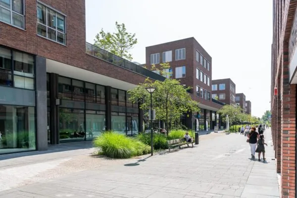 Moderne straat aan de Maasstadweg in Rotterdam met bakstenen gebouwen, winkels, bomen en voetgangers op een zonnige dag.