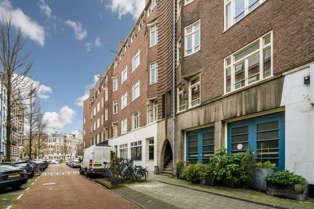 Straatbeeld van de Nassaukade in Amsterdam met karakteristieke bakstenen huizen, geparkeerde auto's en fietsen.