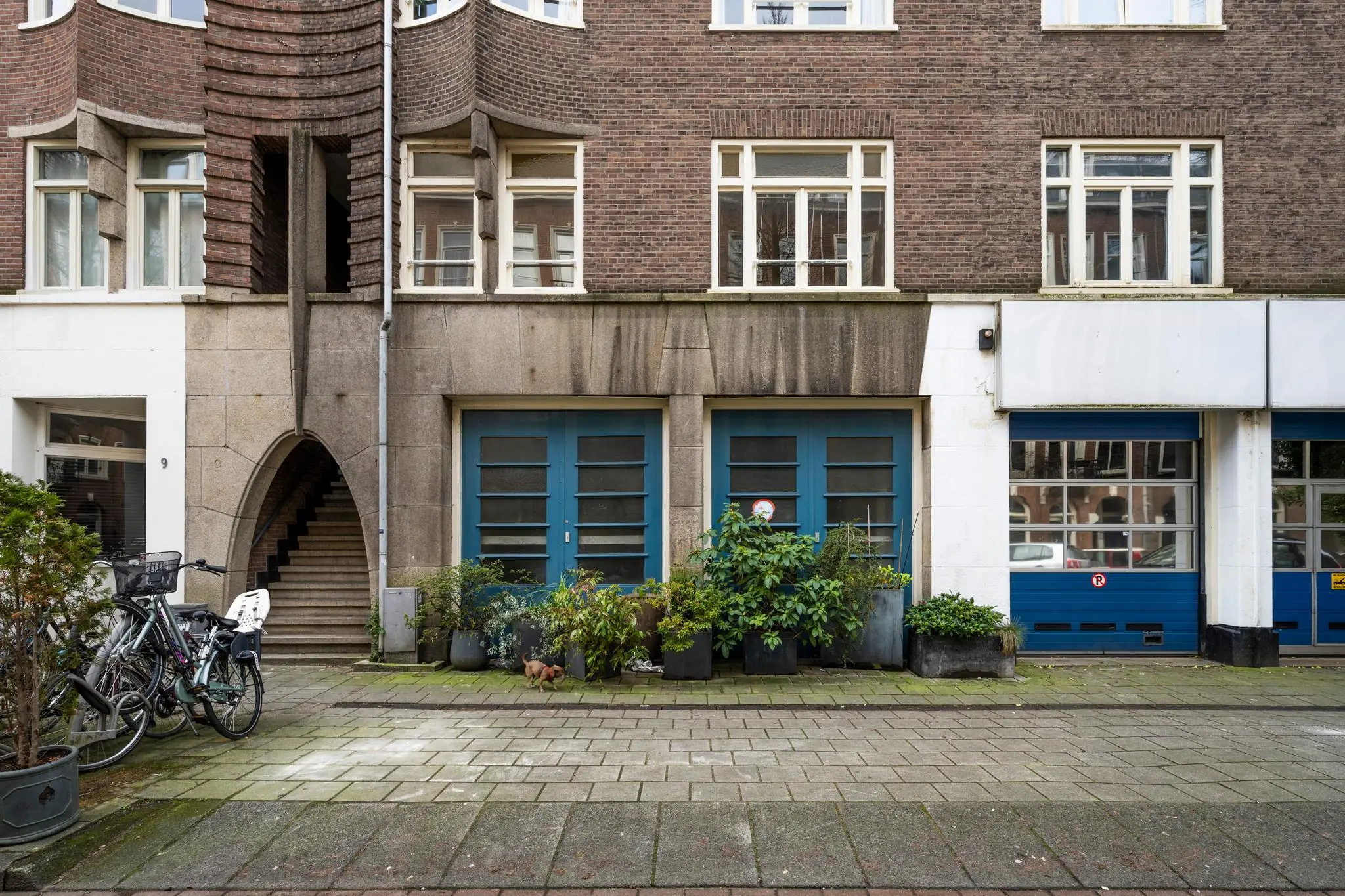 Voorgevel van een woongebouw aan de Nassaukade met blauwe deuren, plantenbakken, fietsen en een trap onder een rondboog.