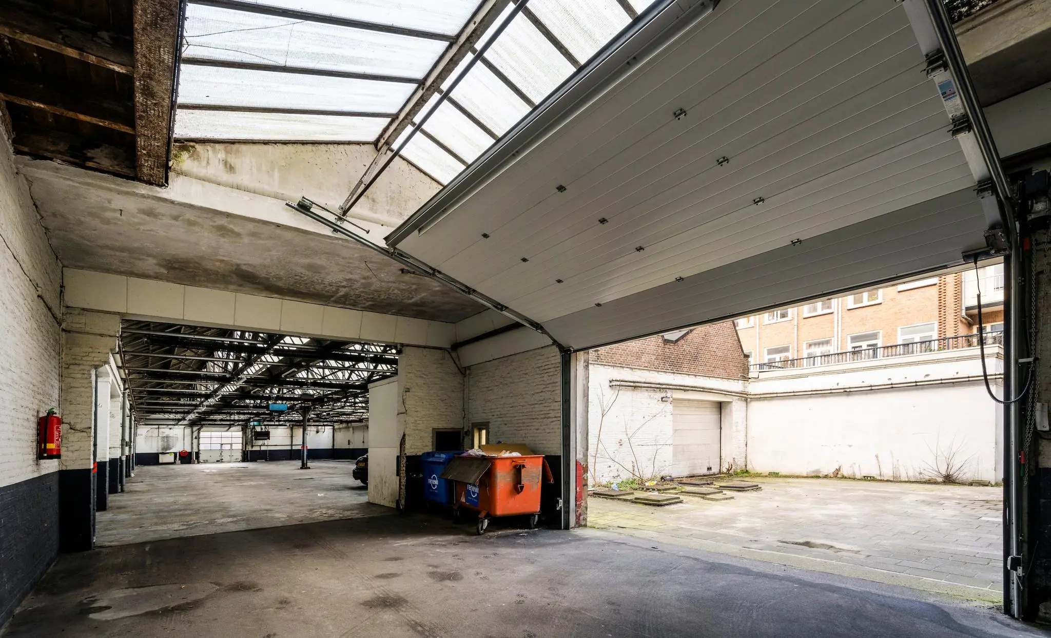 Binnenzicht van een lege loods aan de Nassaukade met openstaande garagedeur en vuilcontainers bij de uitgang.