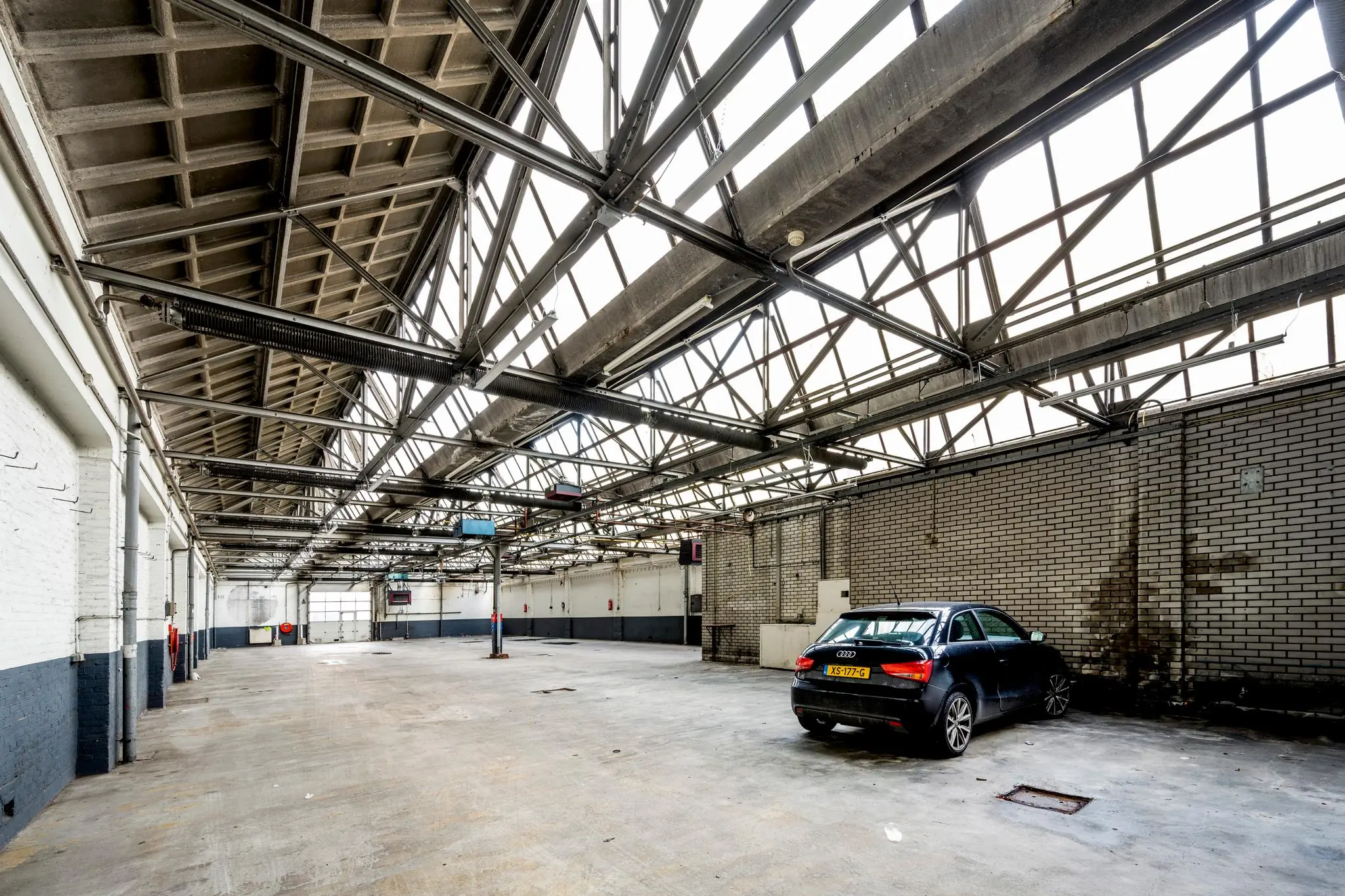 Een lege industriële loods aan de Nassaukade met een geparkeerde zwarte Audi onder een dak met stalen spanten en grote ramen.
