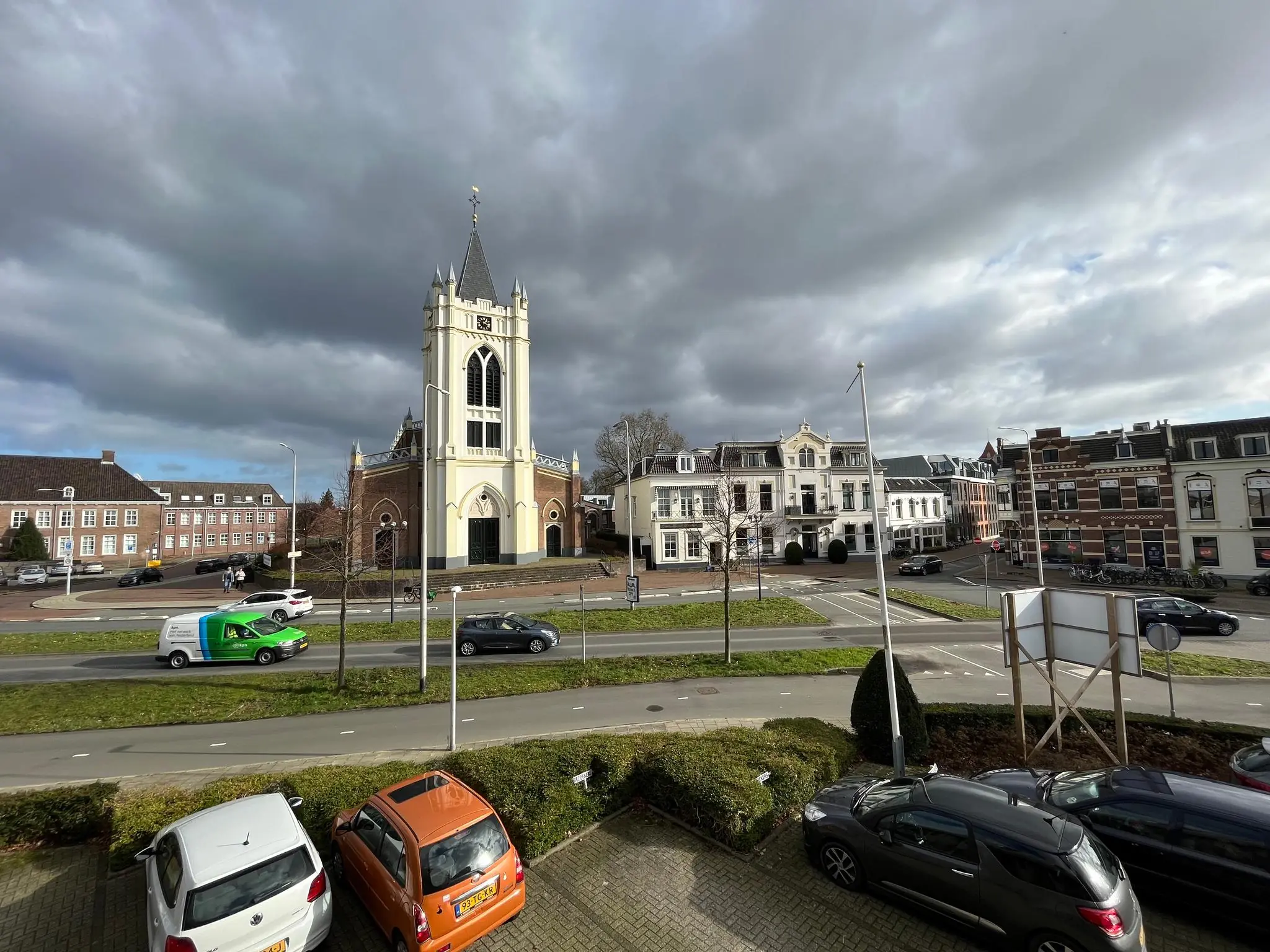 Een straatbeeld van de Utrechtseweg met een neogotische kerk, omliggende historische panden en geparkeerde auto's onder een bewolkte lucht.
