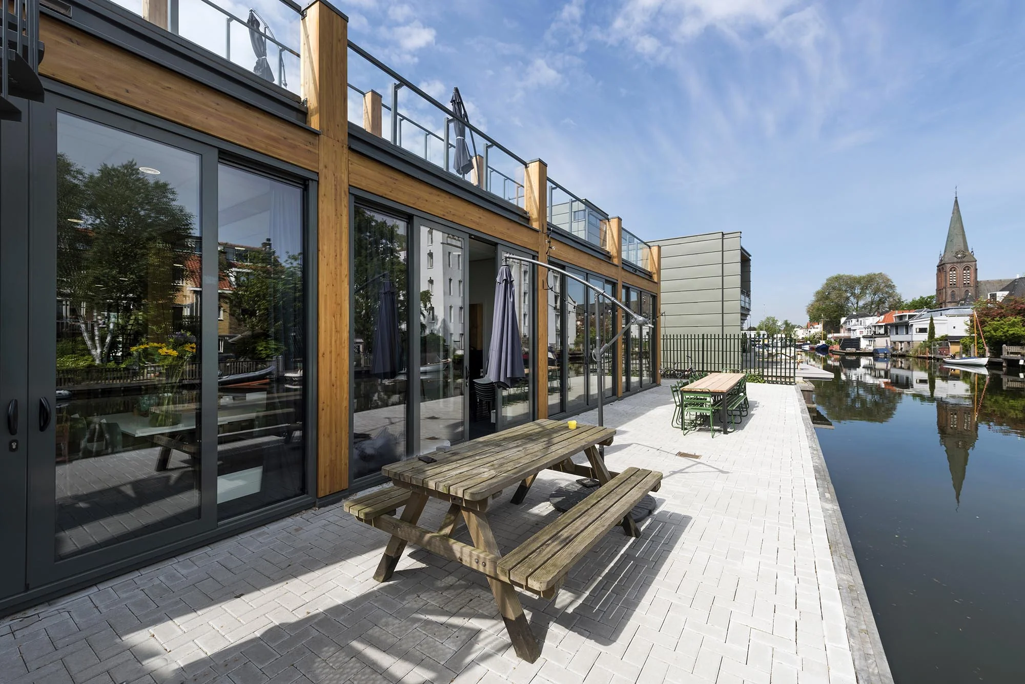 Moderne waterwoningen aan de Nieuwendammerkade met houten gevels, picknicktafels en uitzicht op een gracht en kerktoren.