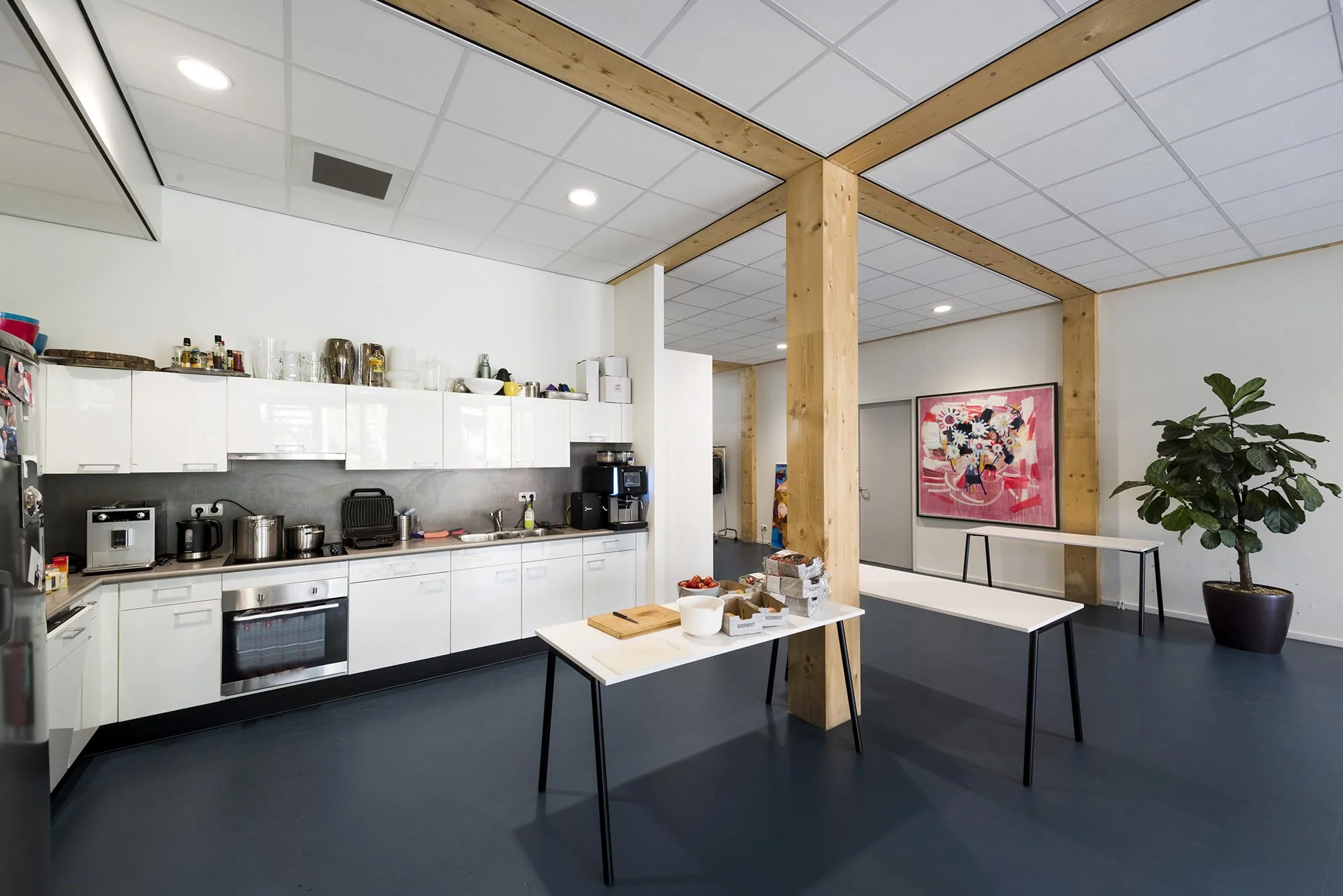 Moderne kantoorkeuken aan de Nieuwendammerkade met witte kasten, houten steunpilaren en een kleurrijk schilderij aan de muur.