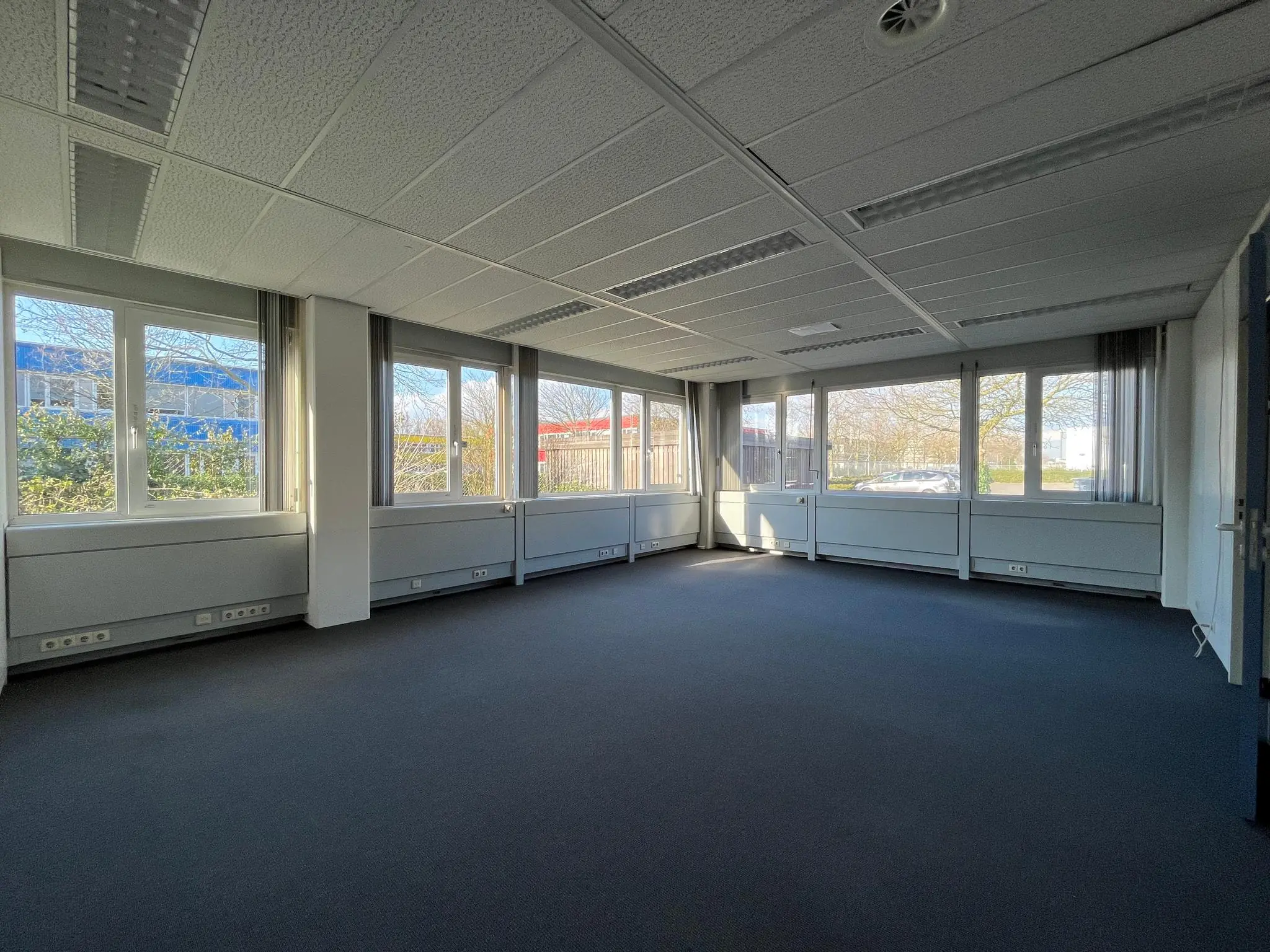 Leeg kantoorruimte met grote ramen aan de Energieweg, met uitzicht op bomen en omliggende gebouwen.