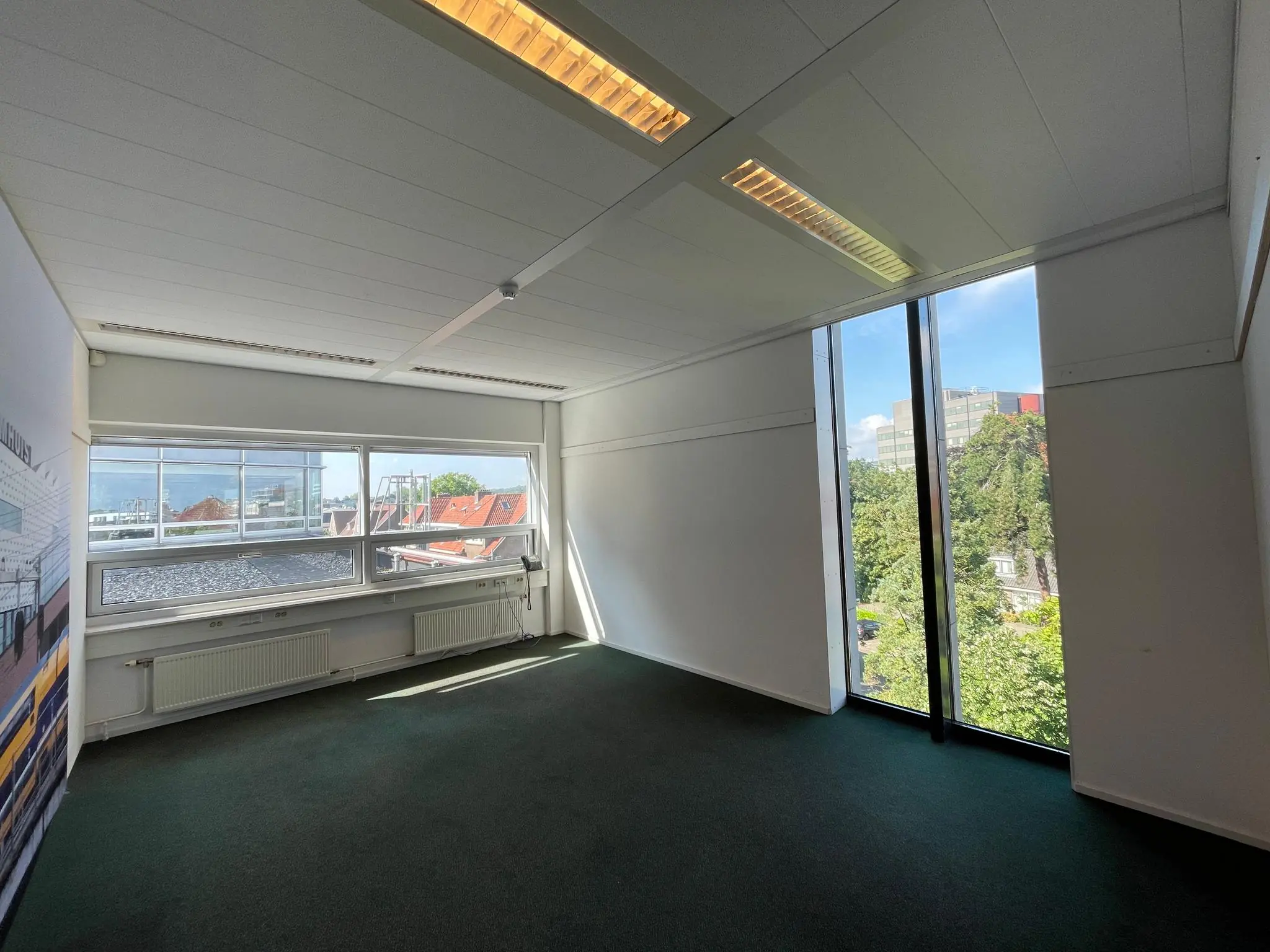 Licht en leeg kantoor met grote ramen aan de Arnhemseweg met uitzicht op daken en bomen.