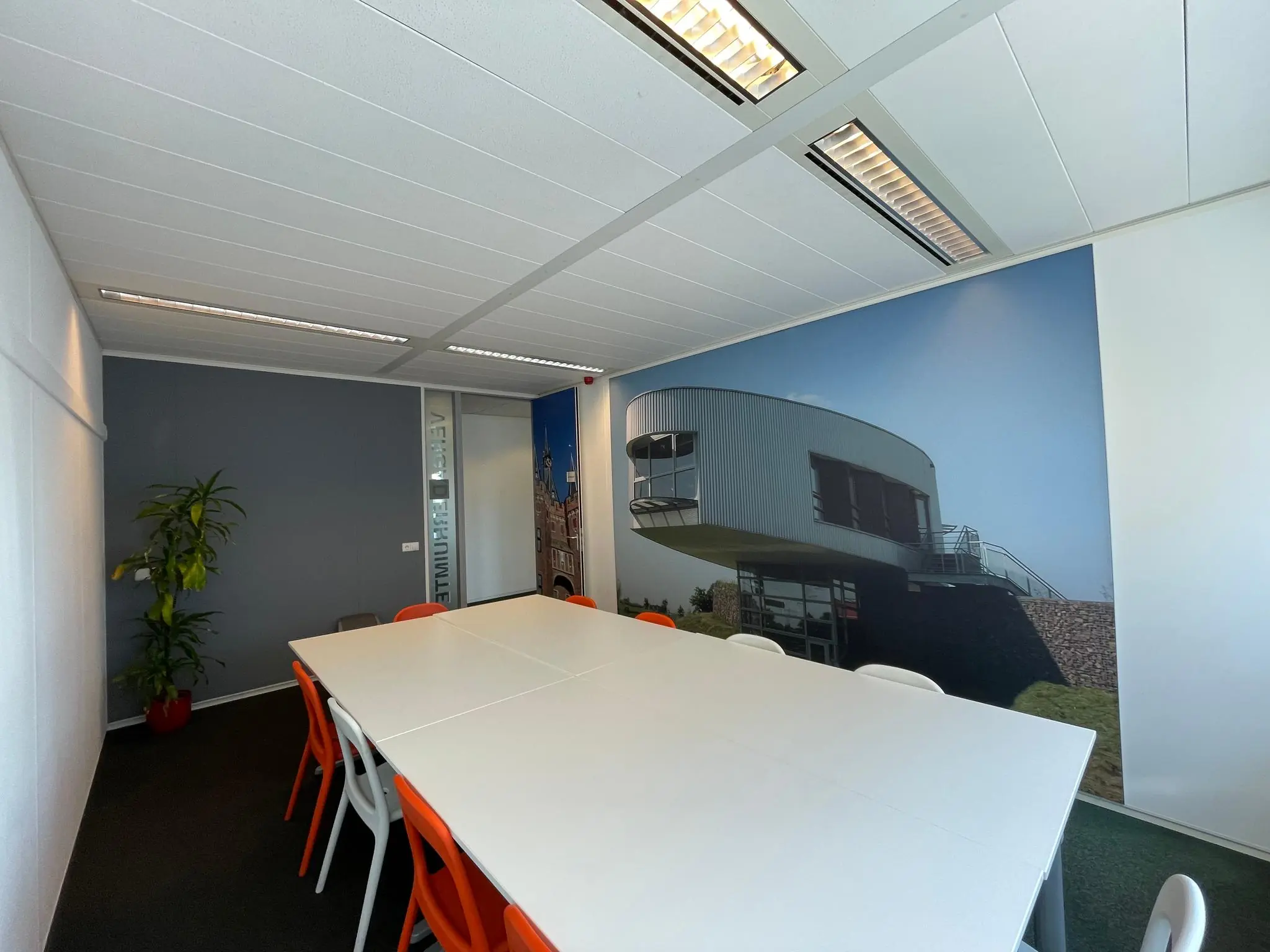Vergaderruimte met witte tafels, oranje en witte stoelen, en een grote wandfoto van een modern gebouw aan de Arnhemseweg.