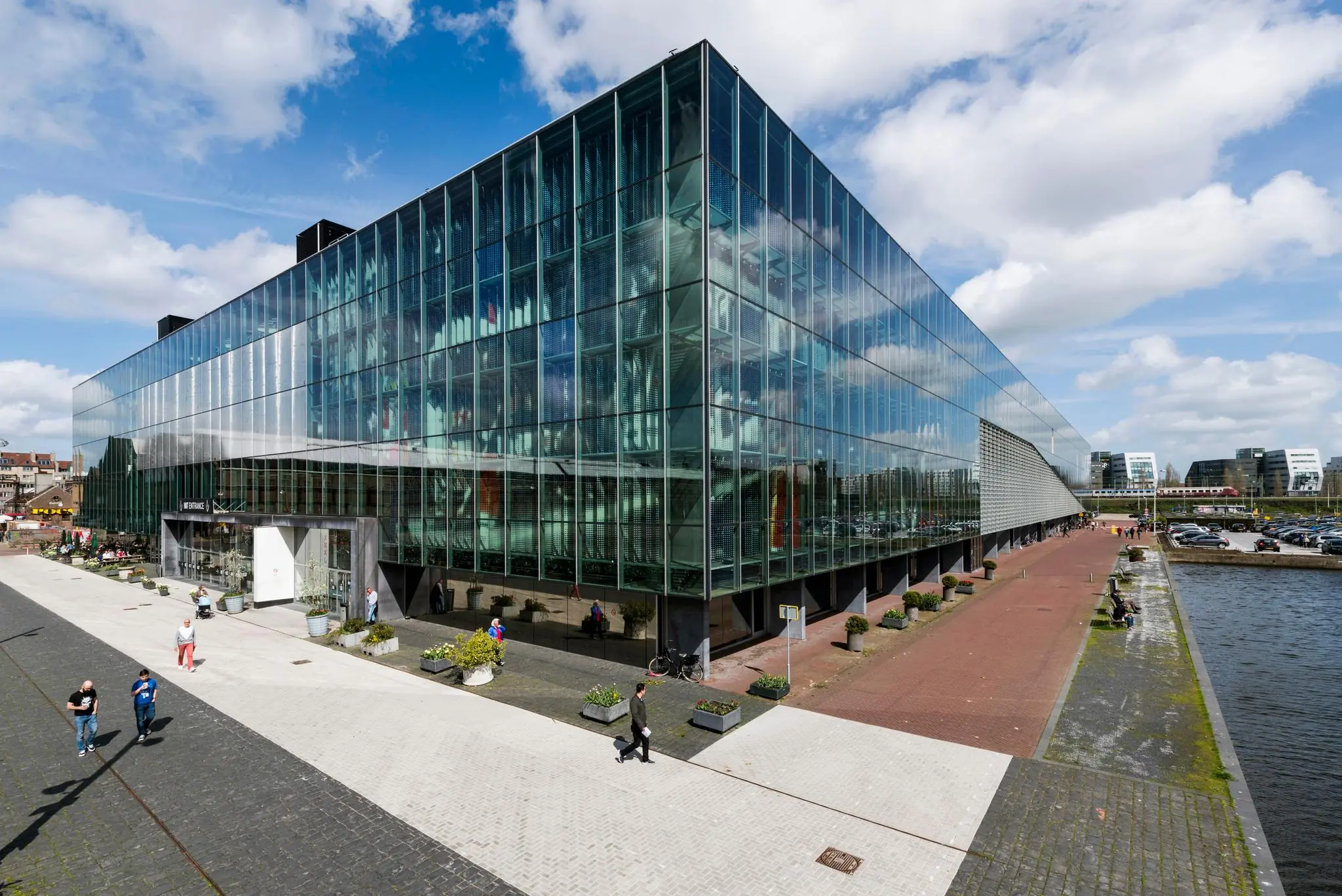 Modern glazen kantoorgebouw aan de Oostenburgermiddenstraat in Amsterdam, gelegen langs een kade met voetgangers en geparkeerde auto's.