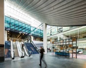 Moderne, lichte binnenruimte aan de Oostenburgermiddenstraat met roltrappen, glazen gevels en een open kantoor- of ontmoetingsruimte.