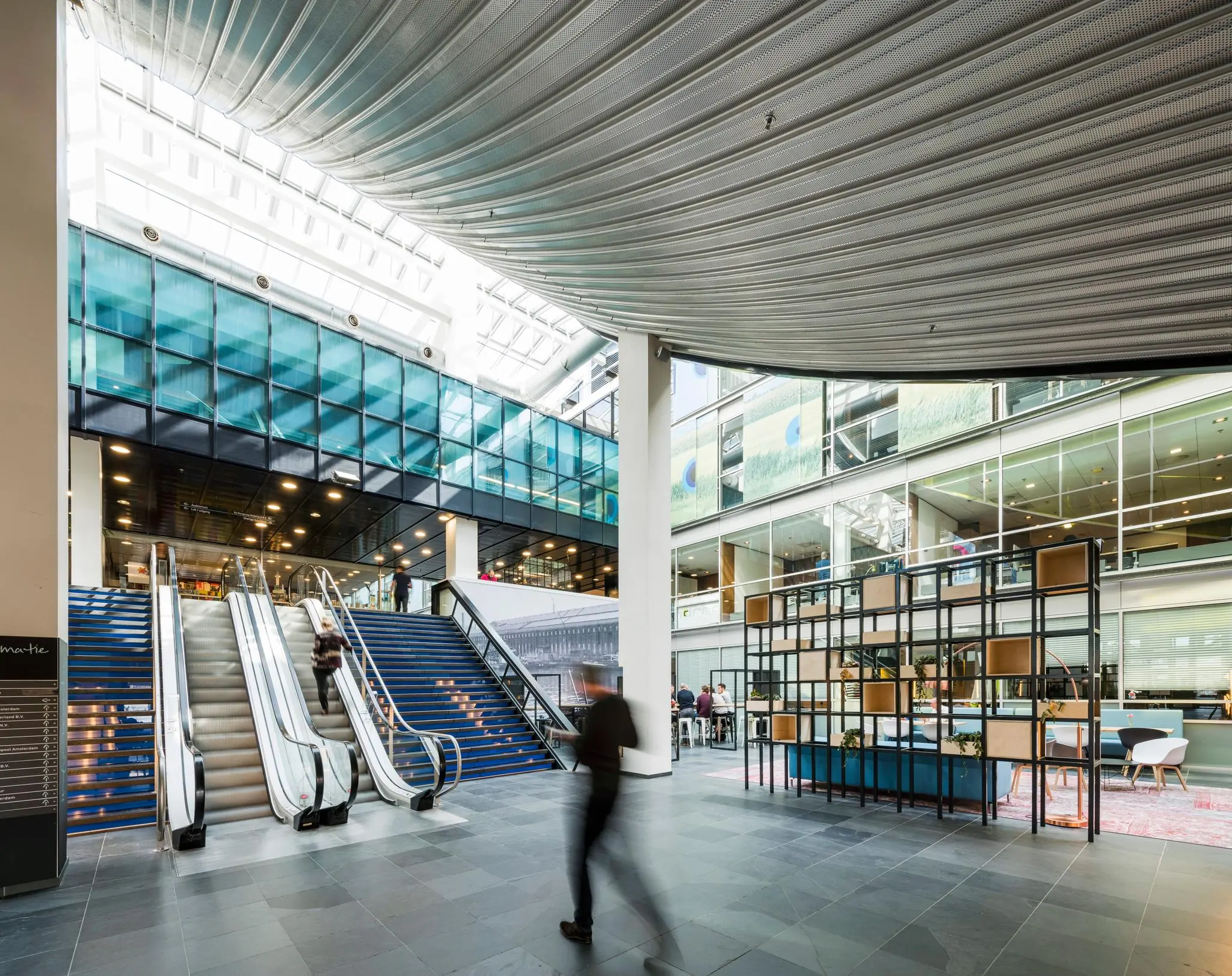 Moderne, lichte binnenruimte aan de Oostenburgermiddenstraat met roltrappen, glazen gevels en een open kantoor- of ontmoetingsruimte.
