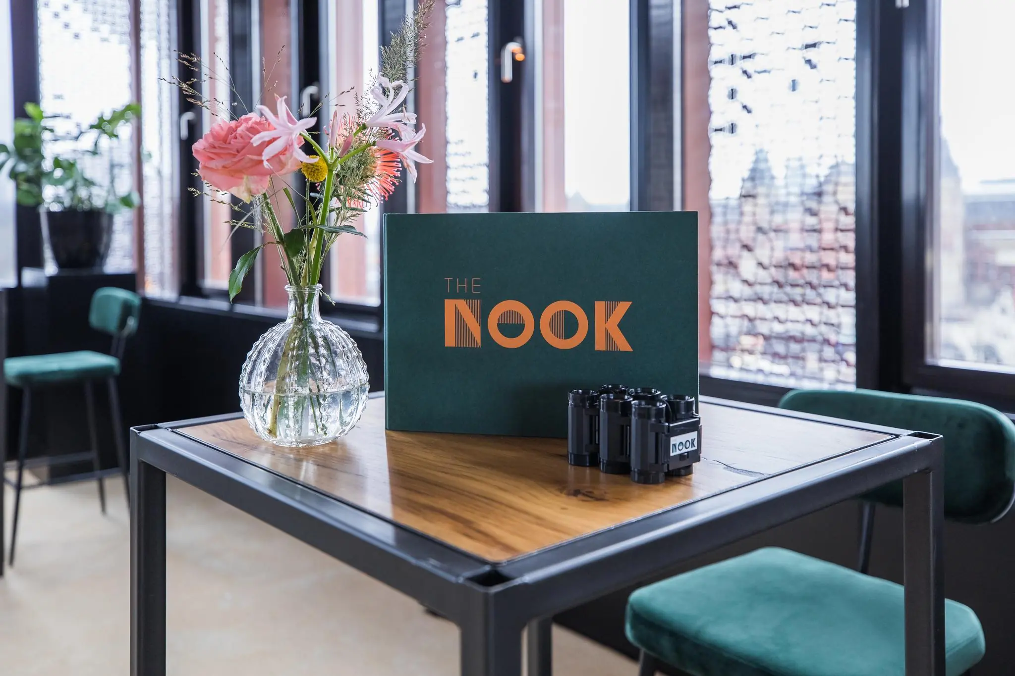 Een houten tafel met een bloemenvaas en een bord met het logo van "The Nook" in een modern interieur aan de Prins Hendrikkade.