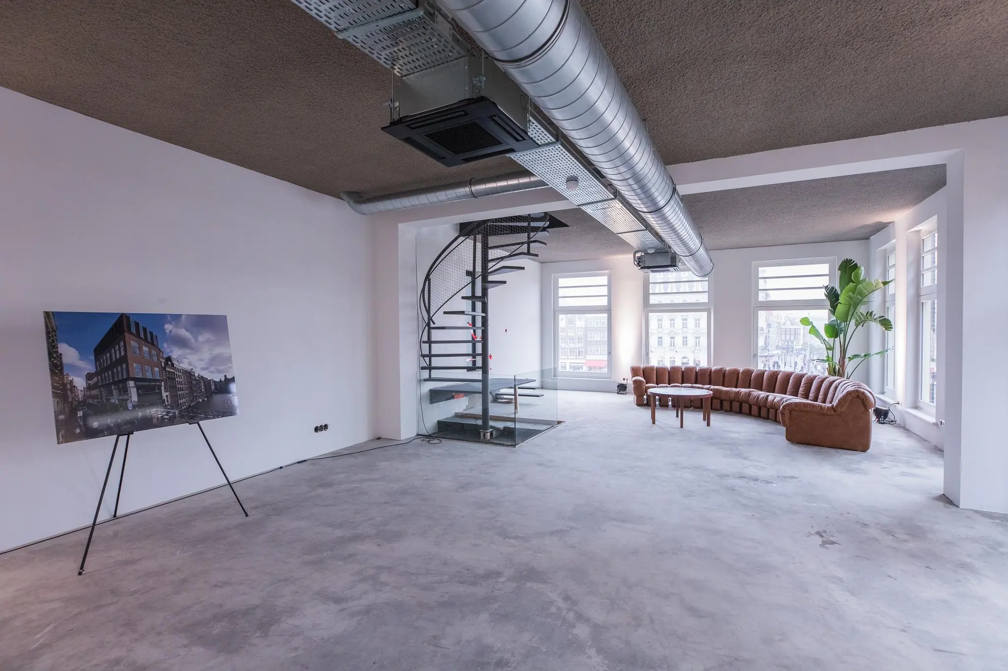 Industriële loft aan de Prins Hendrikkade met een spiraaltrap, grote ramen, bruine hoekbank en poster van het gebouw.