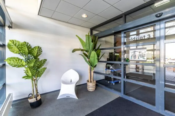Moderne entree van gebouw Veldzigt 3 met glazen schuifdeur, witte designstoel en grote kamerplanten.
