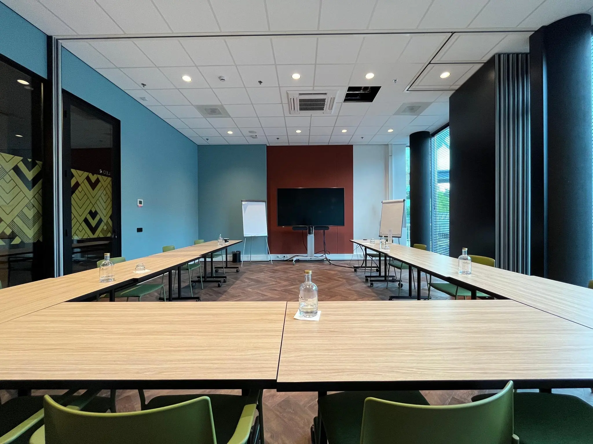 Vergaderruimte op het Herculesplein met U-vormige opstelling, presentatiescherm, flip-overs en flessen water op tafel.