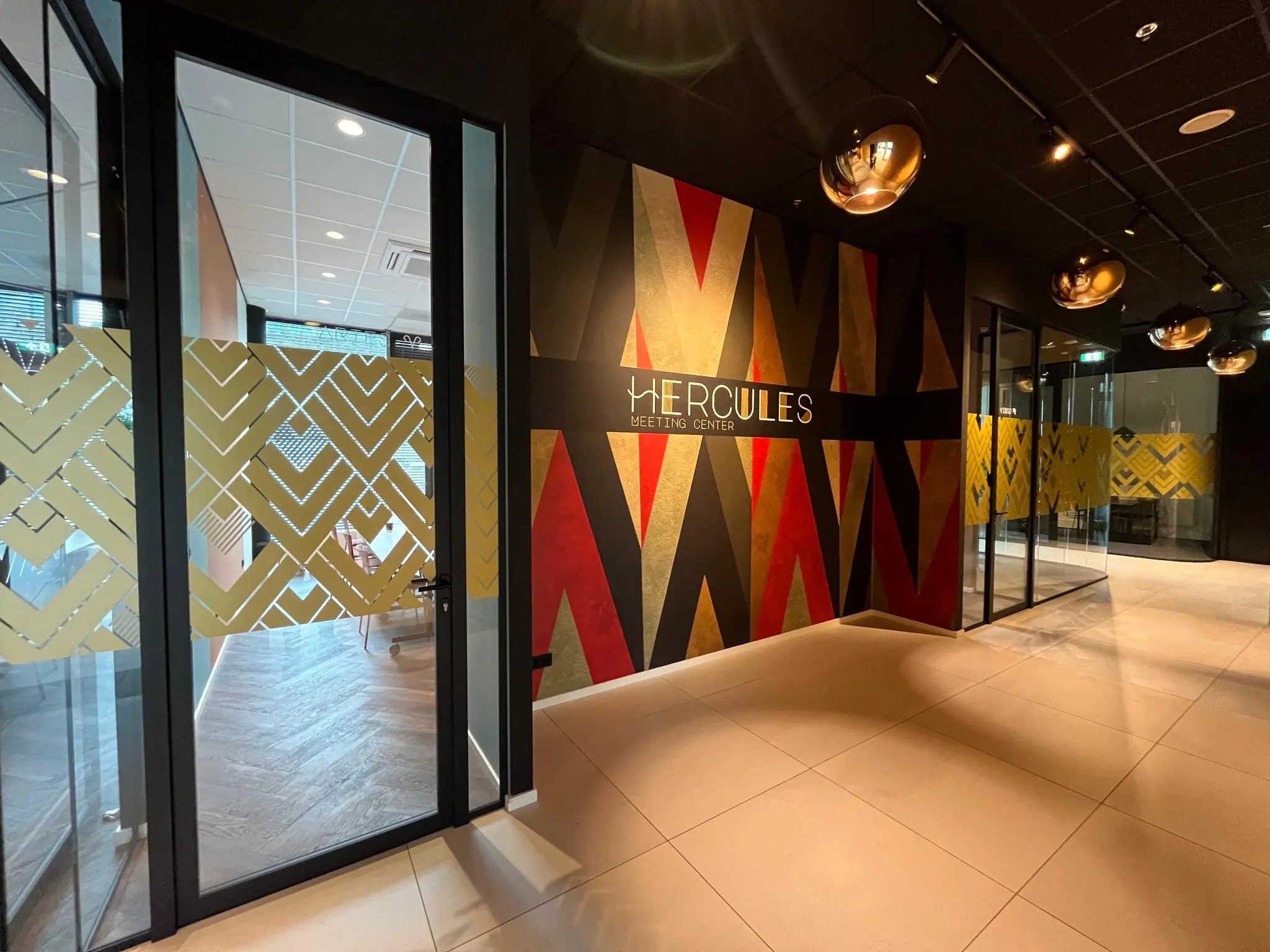 Ingang van het Hercules Meeting Center met moderne glazen wanden en een kleurrijke, geometrisch gedecoreerde muur.