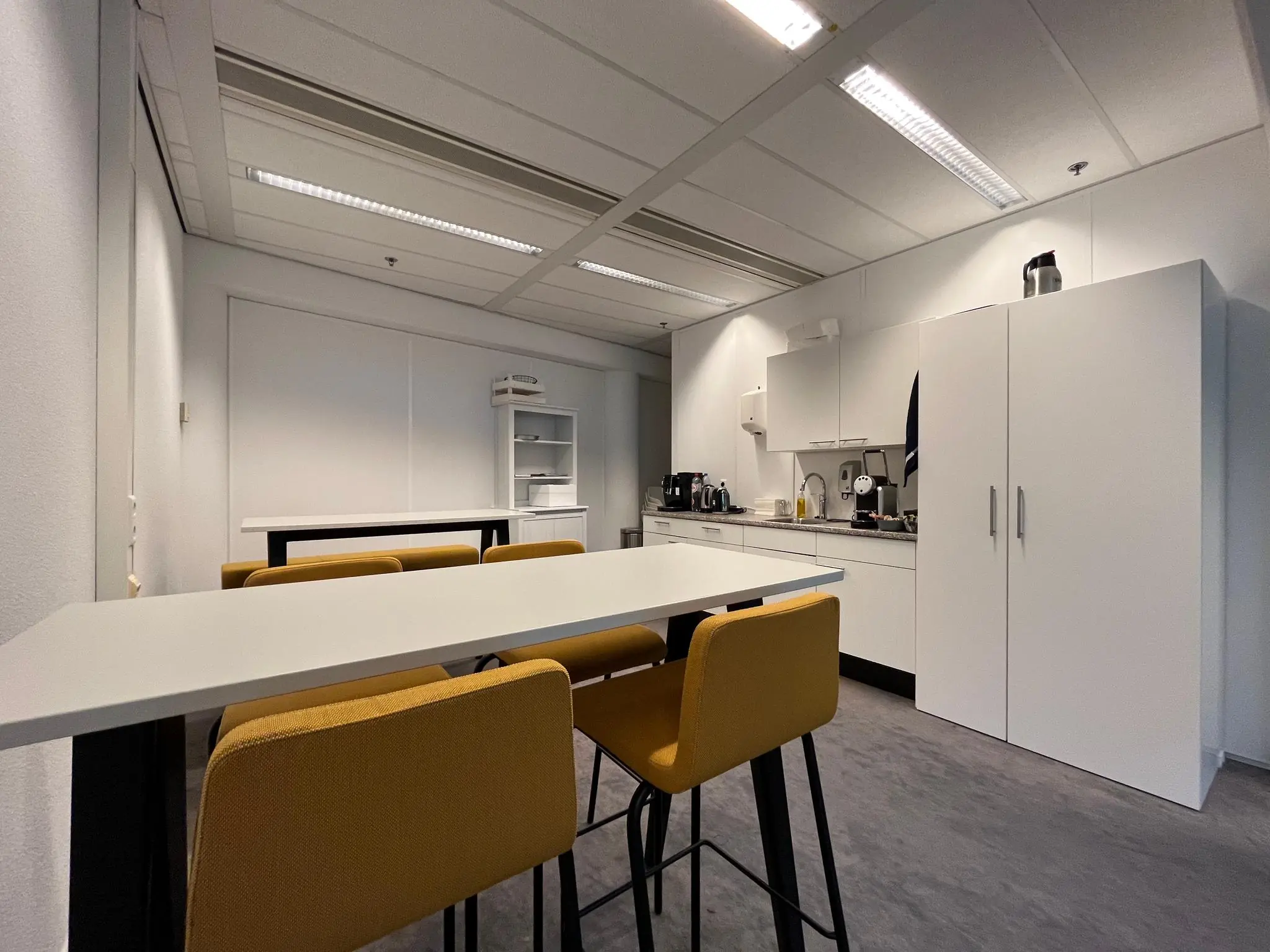 Moderne kantoorpantry met witte keukenkasten, een hoge tafel en gele barkrukken op Herculesplein.