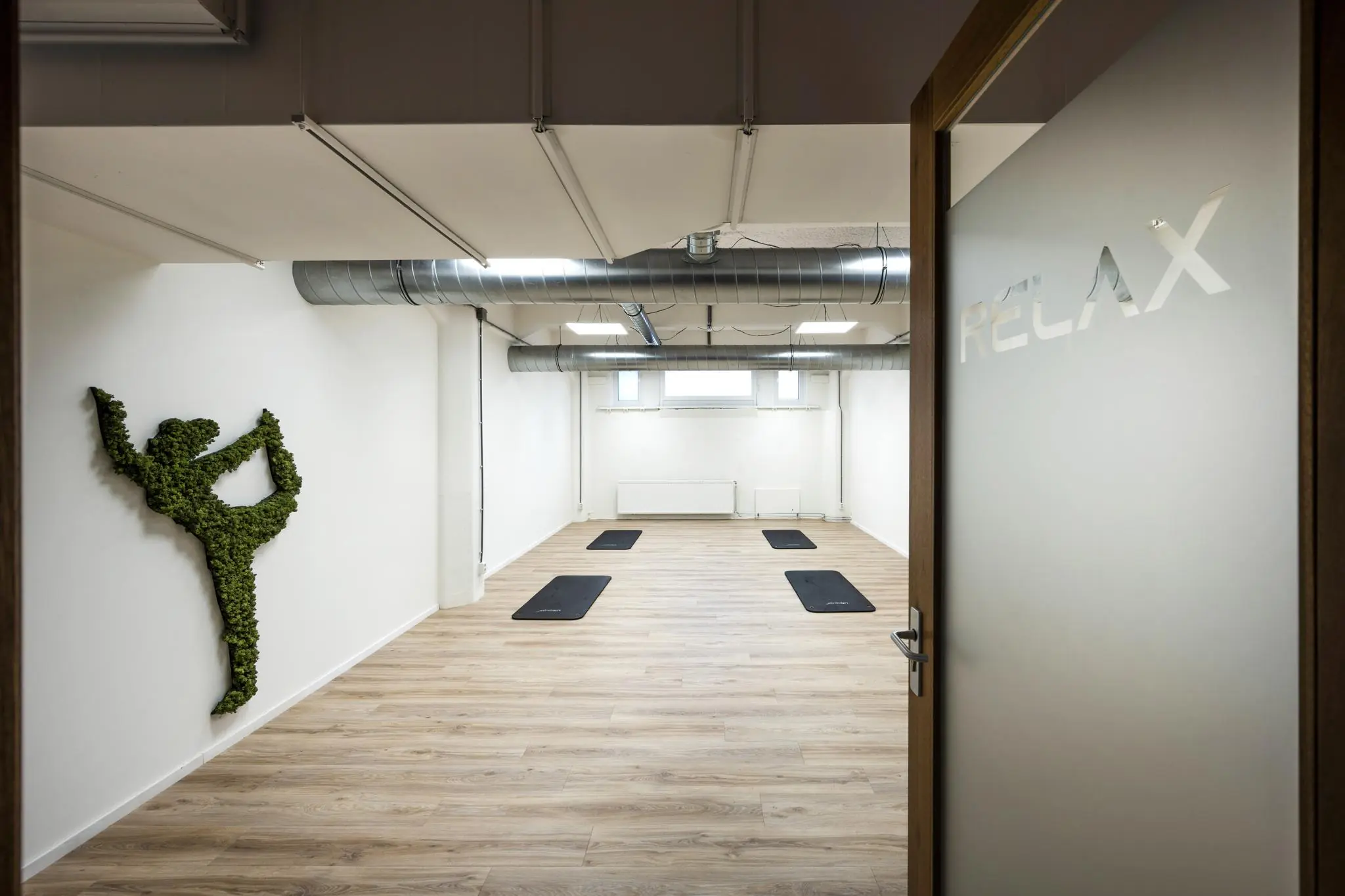 Minimalistische yogastudio aan de Van Diemenstraat met yogamatten op een houten vloer, een muurdecoratie van mos in yogahouding en een deur met het woord "RELAX".