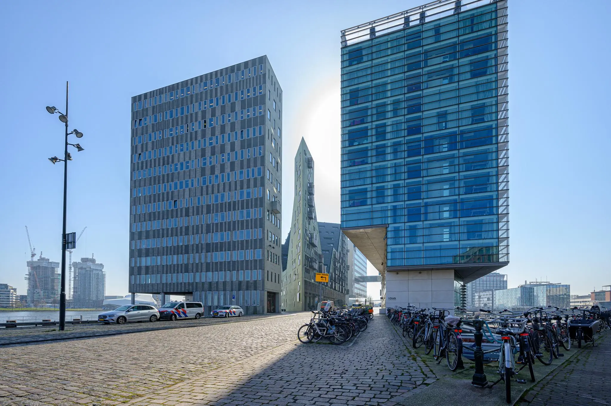 Straatbeeld bij het IJdok in Amsterdam met moderne kantoorgebouwen, geparkeerde fietsen en politiewagens.