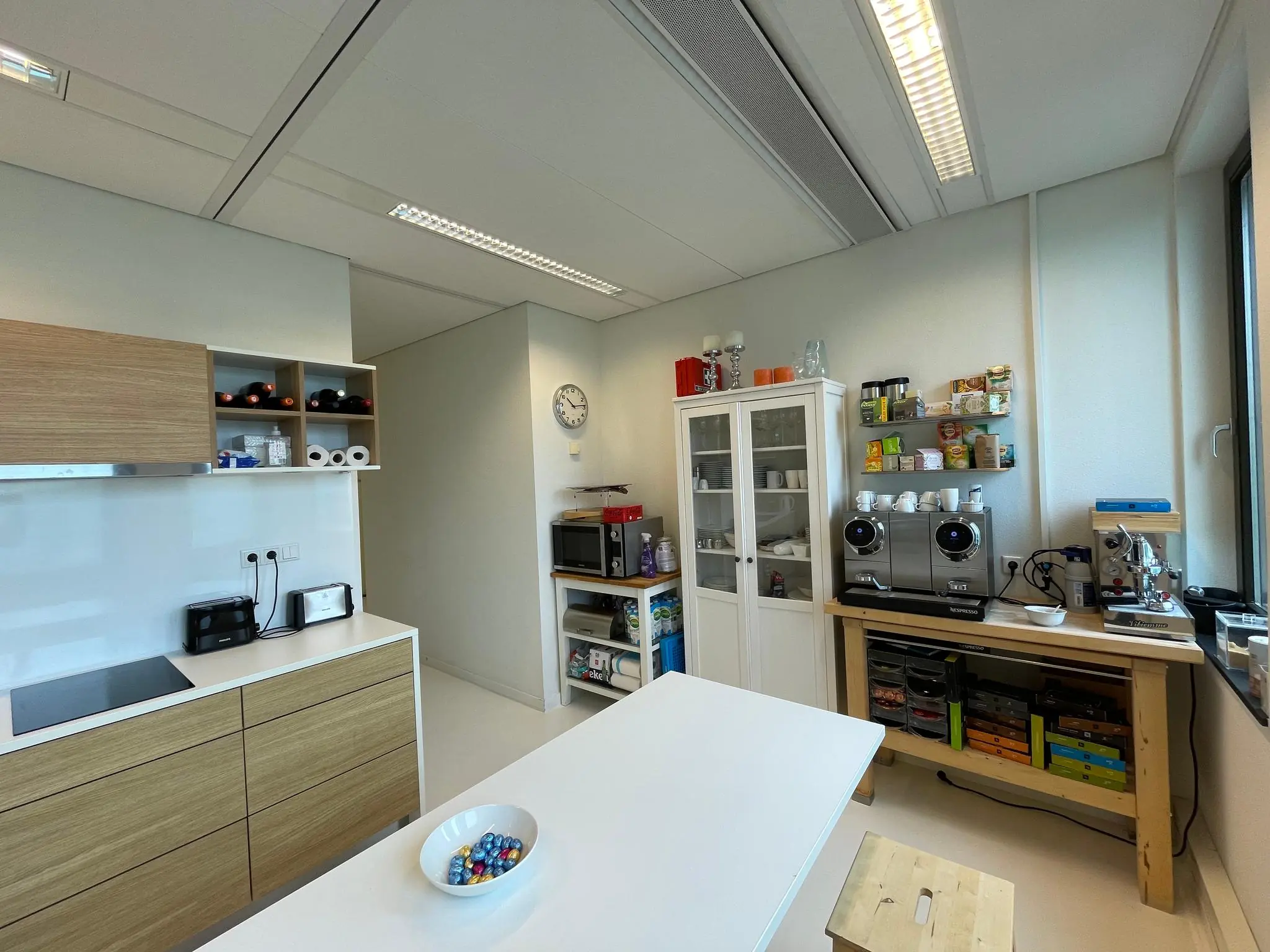 Moderne kantoorkeuken met koffiezetapparaten, voorraadkast en een centraal wit werkblad.