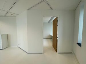 Kantoorruimte met witte wanden en vloer, kijkend naar een openstaande houten deur die leidt naar een aangrenzend vertrek.