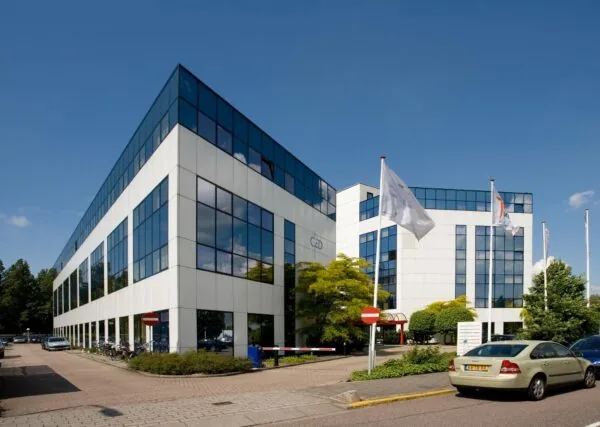 Modern kantoorgebouw aan de Rietbaan met witte gevel en spiegelende ramen, omgeven door vlaggen, auto's en groen.