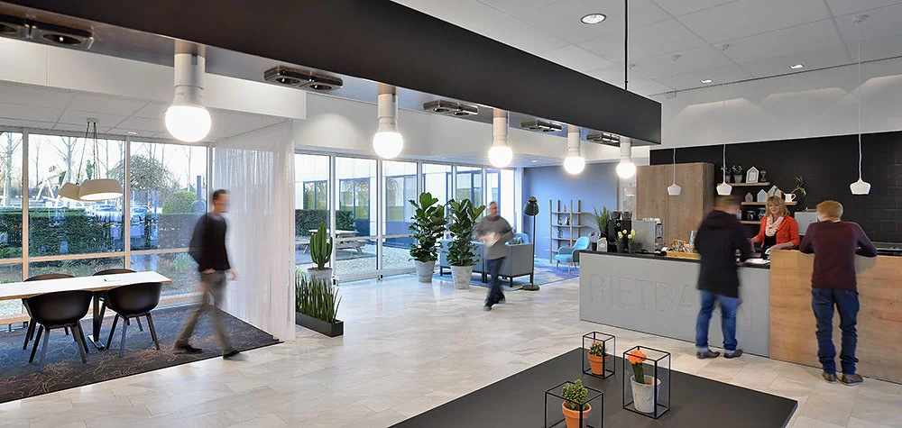 Moderne, lichte ontvangsthal van kantoorpand Rietbaan met balie, zitgedeelte en enkele bezoekers in beweging.