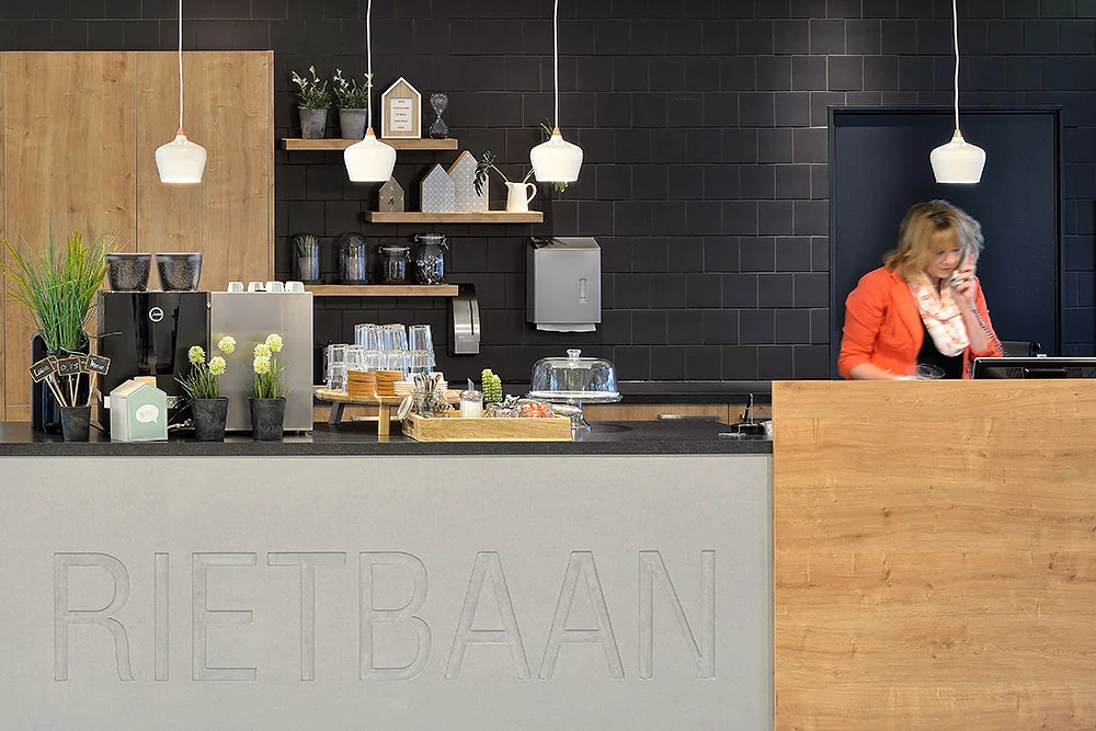 Moderne receptiebalie met koffiecorner bij Rietbaan, waar een vrouw telefoneert achter de balie.
