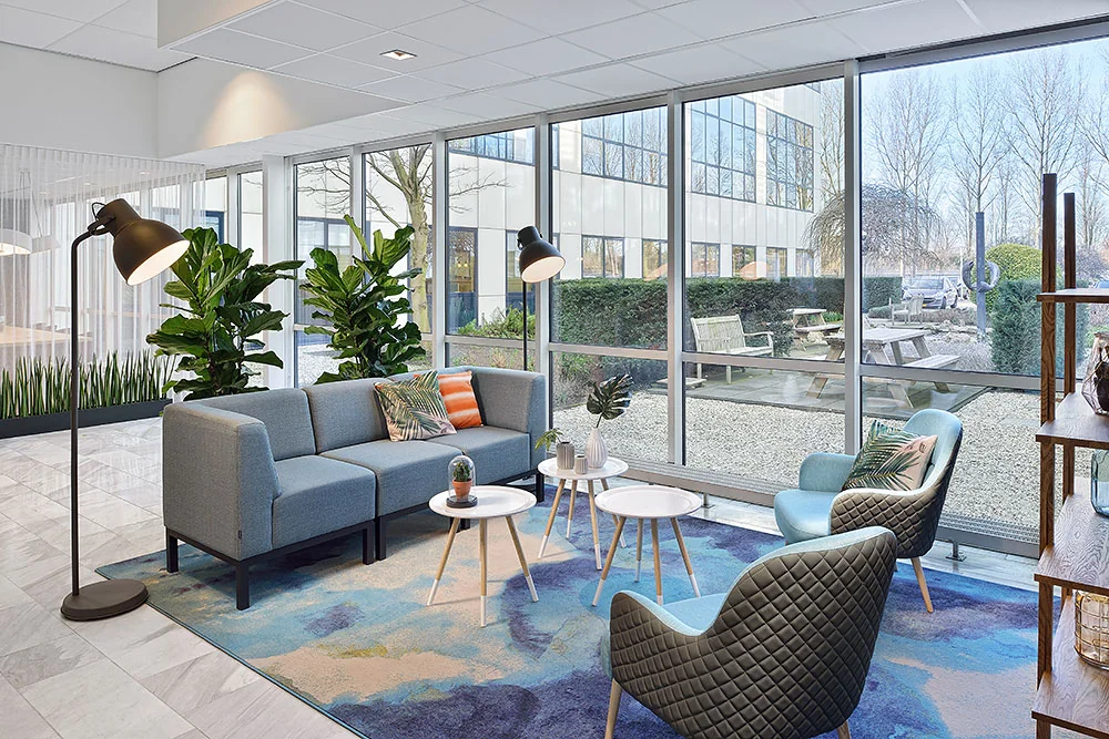 Modern ingerichte wachtruimte op de Rietbaan met comfortabele zitplaatsen en uitzicht op een buitenruimte met picknicktafels.