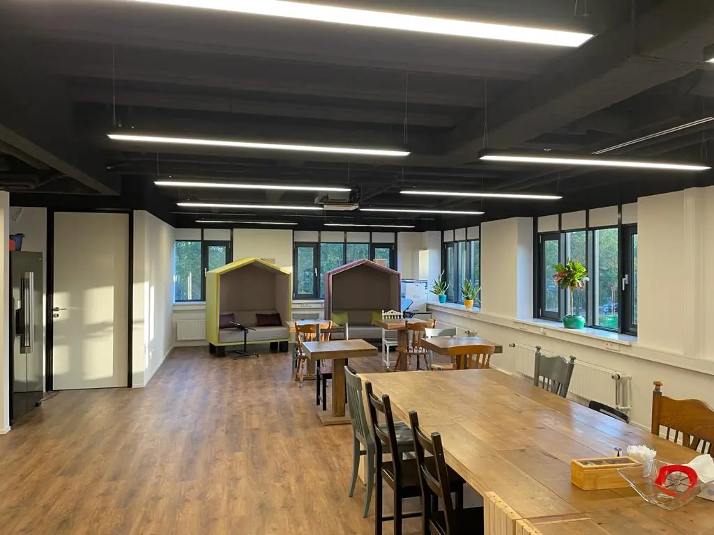 Moderne gemeenschappelijke ruimte op de Rietbaan met houten vloer, lange tafel, huisvormige zithoeken en veel natuurlijk licht.