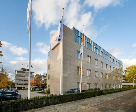 Kantoorgebouw van ICS Groep met vlaggenmasten op de voorgrond en een parkeerplaats aan de zijkant.
