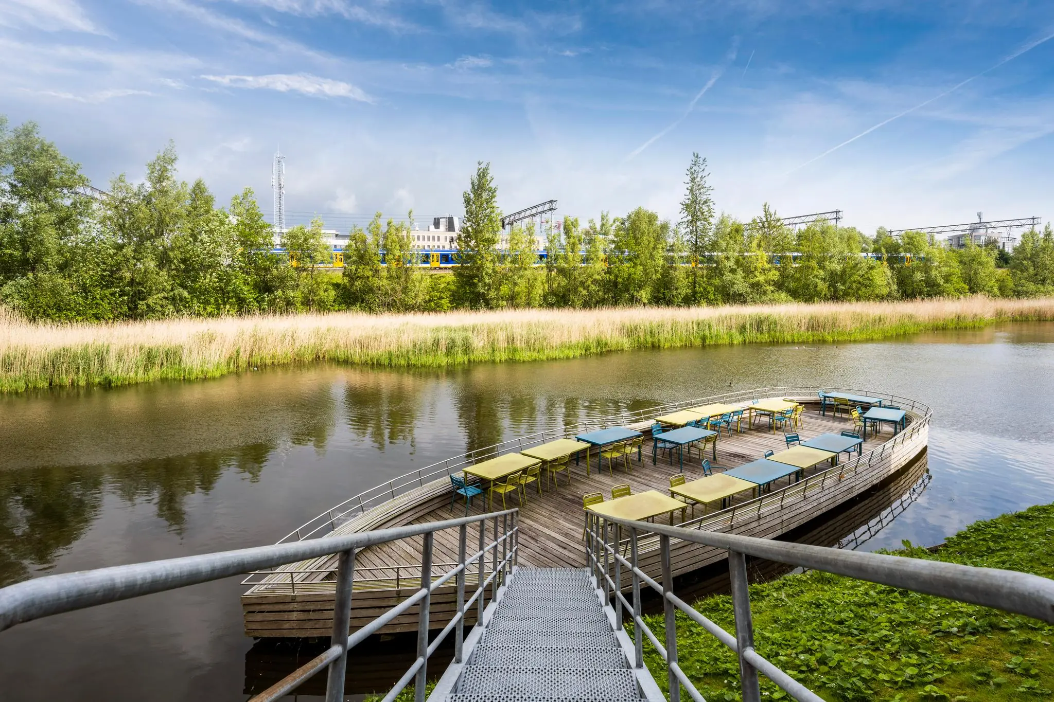 Vlonderterras met kleurrijke tafels en stoelen aan het water, met op de achtergrond de Capellalaan en een passerende trein.