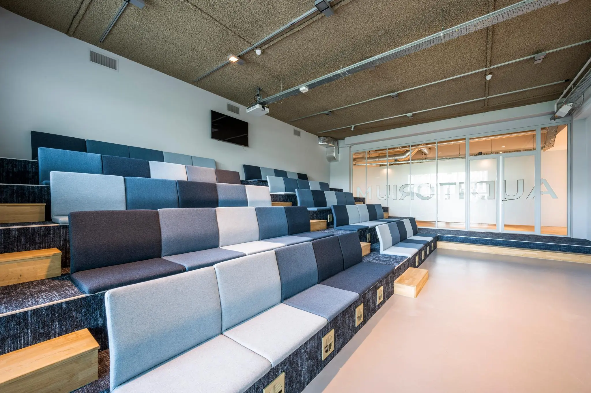 Moderne, kleurrijke tribune-opstelling in een auditorium aan de Capellalaan met gestoffeerde zitplaatsen en houten treden.