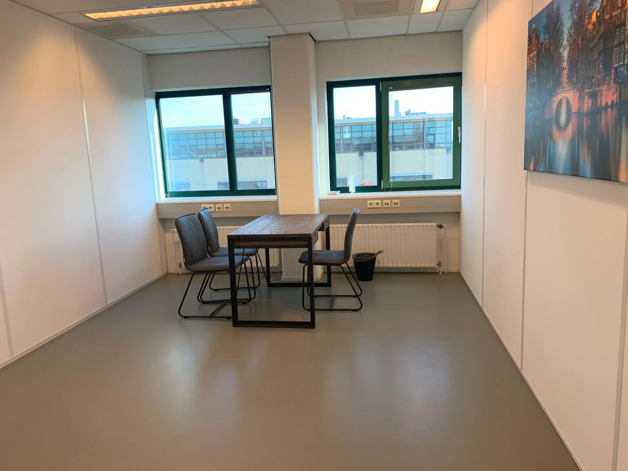 Kleine vergaderruimte aan de Zekeringstraat met een houten tafel, vier stoelen en twee ramen met uitzicht op een kantoorgebouw.