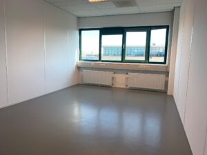 Leeg kantoorruimte met witte wanden, een grijze vloer en een raam met uitzicht op een bedrijventerrein aan de Zekeringstraat.