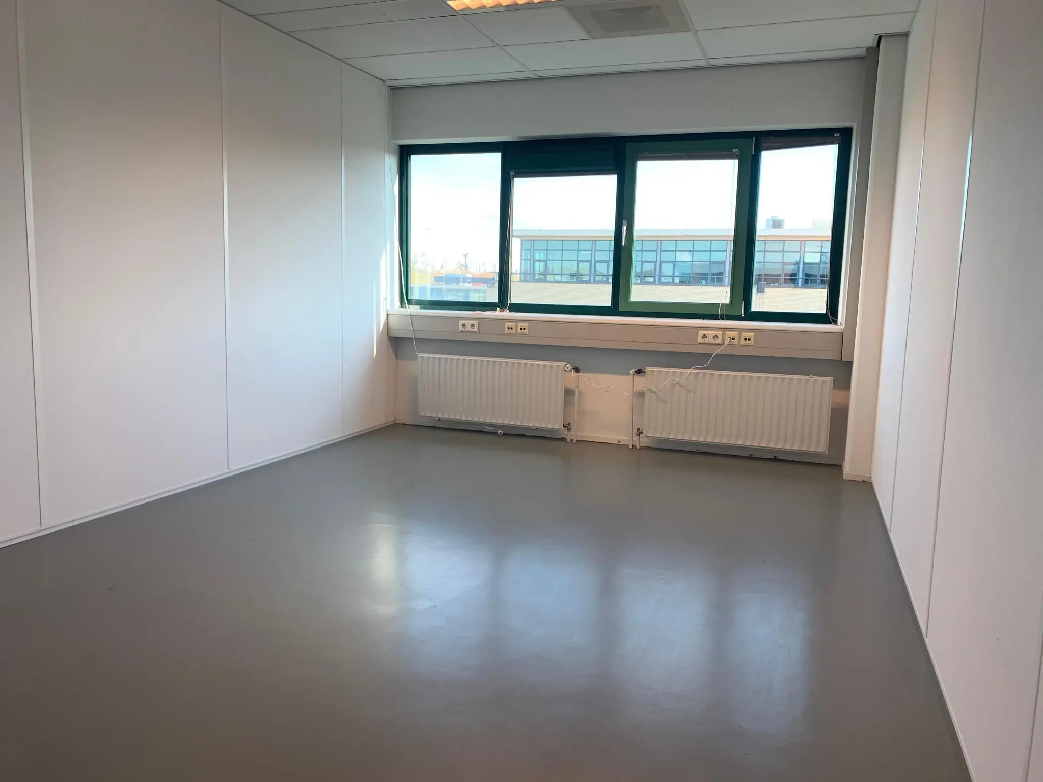 Leeg kantoorruimte met witte wanden, een grijze vloer en een raam met uitzicht op een bedrijventerrein aan de Zekeringstraat.