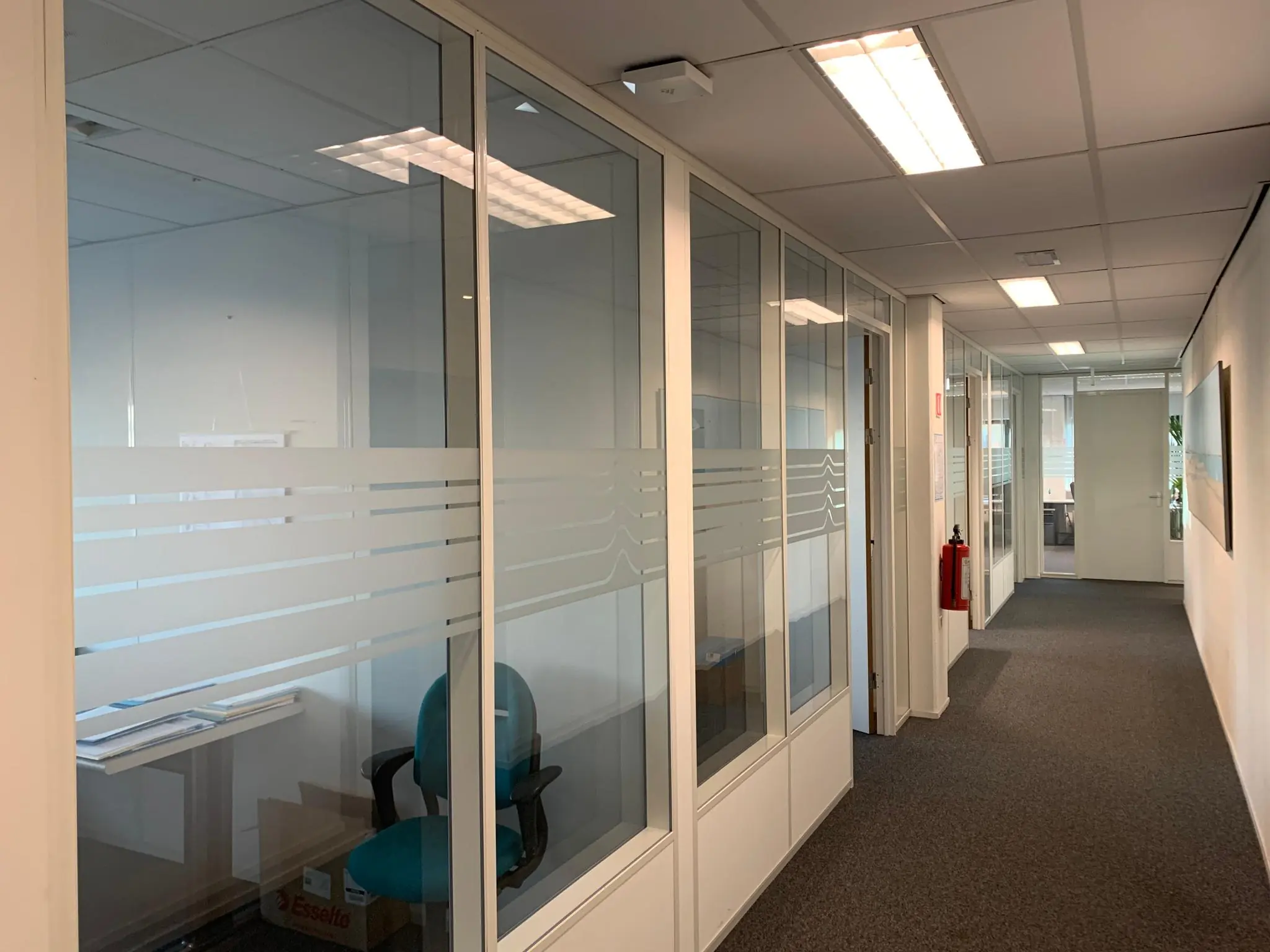 Kantoorgang met glazen vergaderruimtes aan de Zekeringstraat, voorzien van bureaus, stoelen en tl-verlichting.