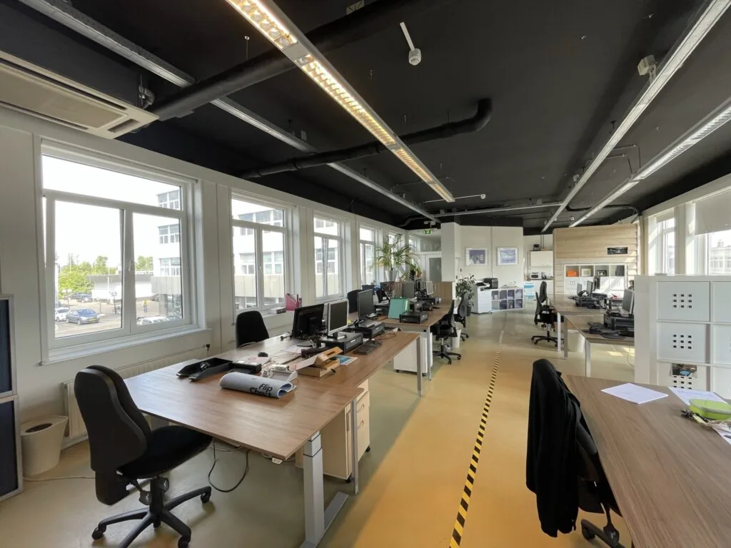 Modern kantoorpand aan de Pilotenstraat met meerdere bureaus, computers en grote ramen die veel daglicht binnenlaten.