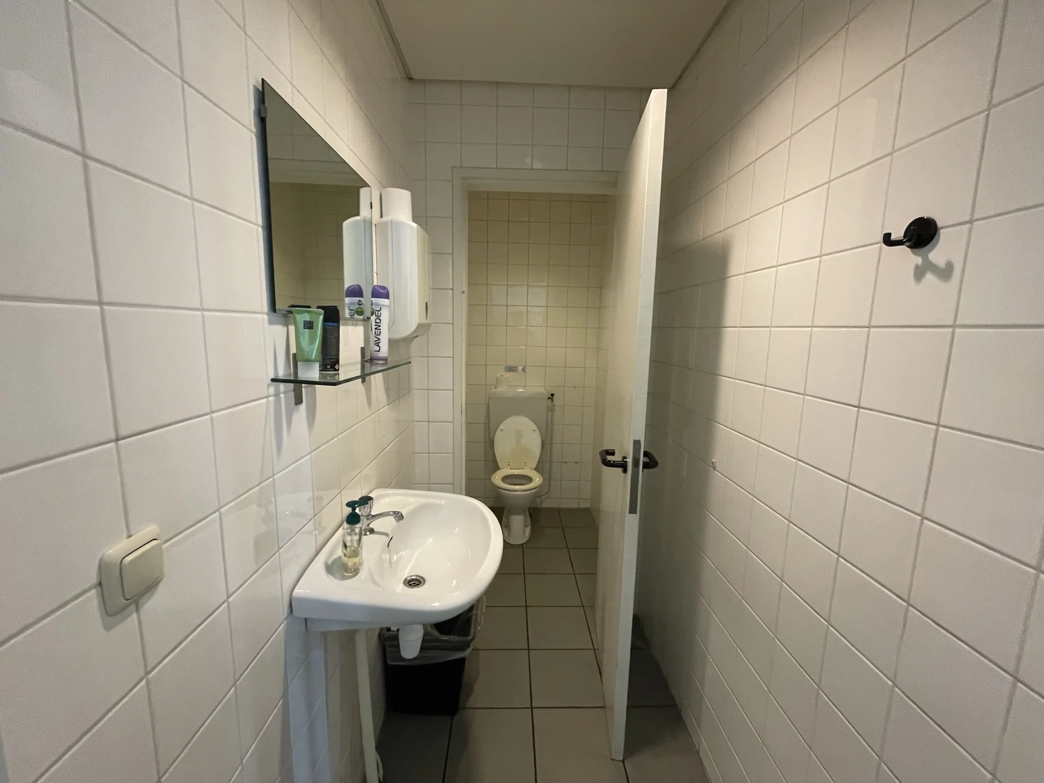 Wit betegelde toiletruimte met een wastafel, spiegel, en een open deur naar het toilet.