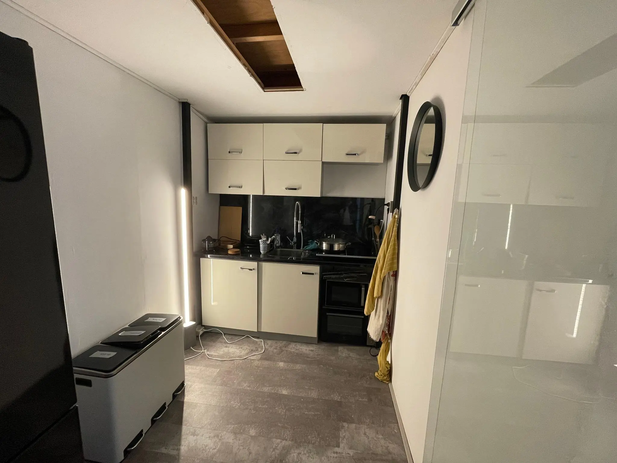 Kleine keuken aan de Nassaukade met beige kastjes, zwarte apparatuur en een plafondopening.