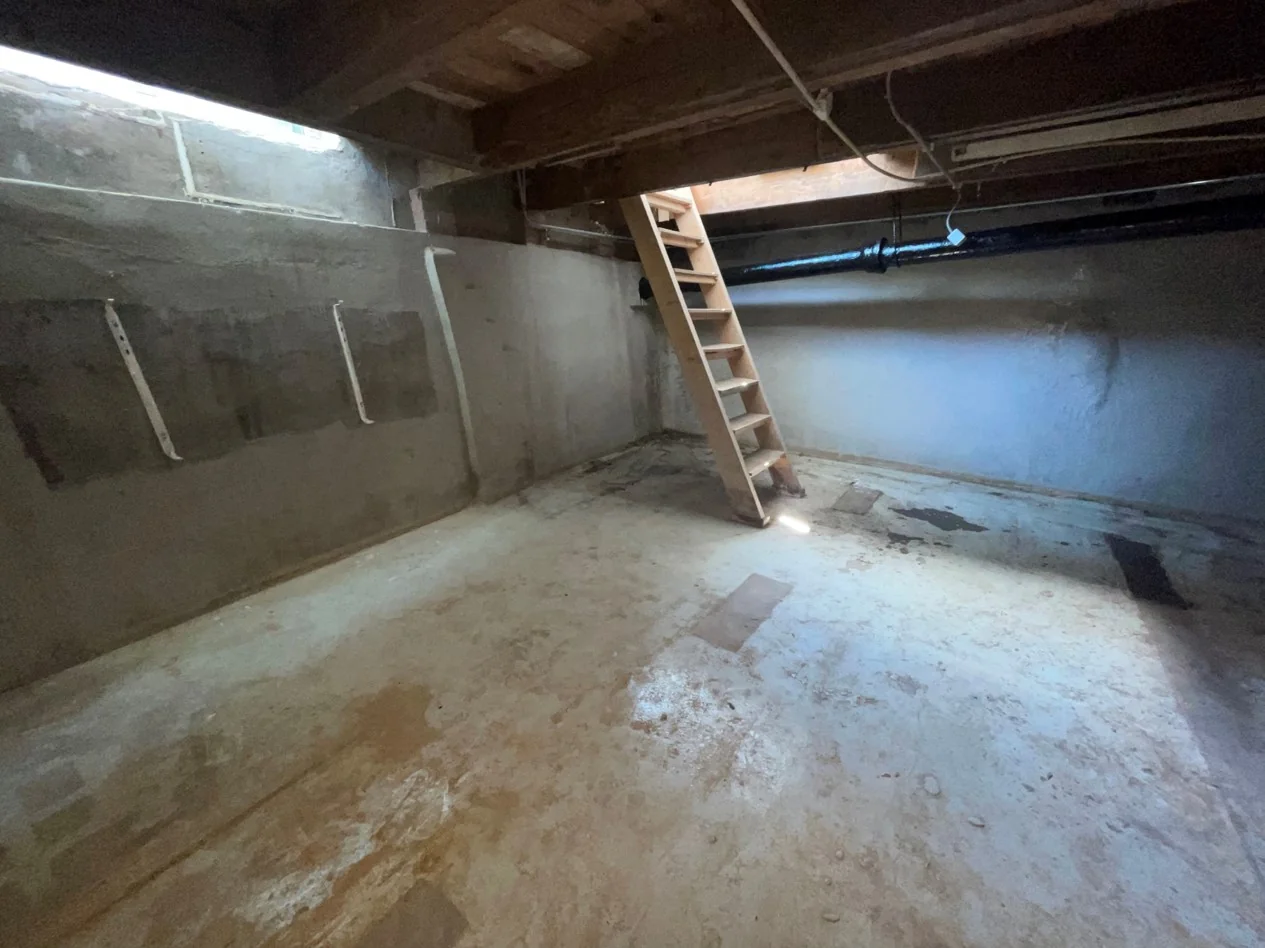 Kelderruimte met betonnen vloer, houten trap en zichtbare leidingen aan het plafond aan de Nassaukade.