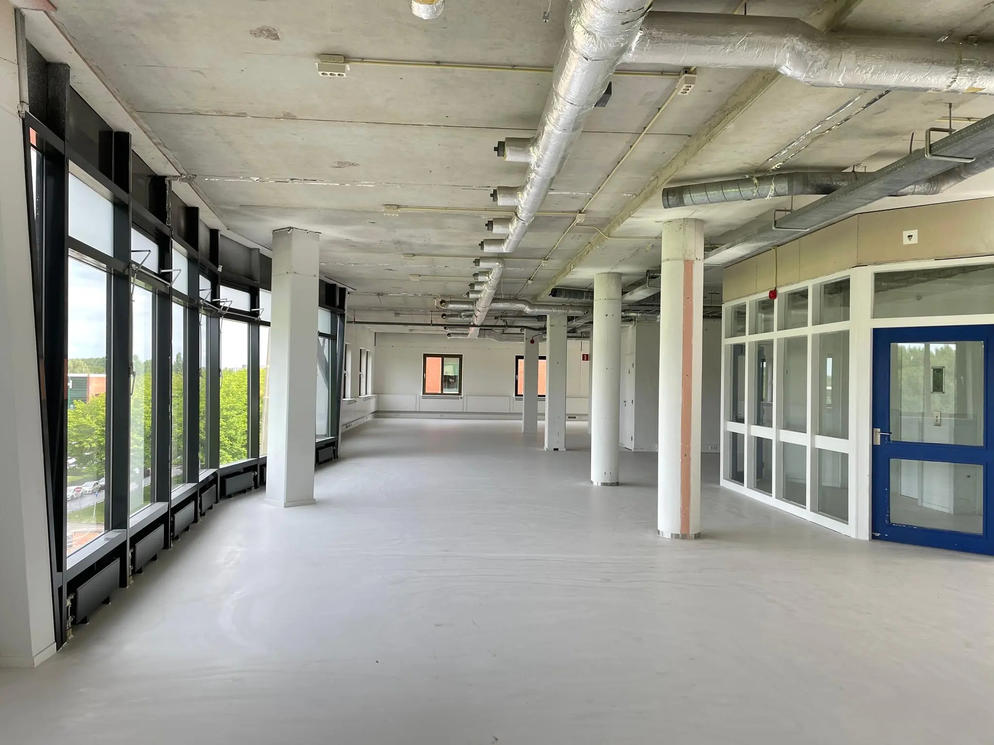 Leeg kantoorinterieur aan de Lichtenauerlaan met grote ramen, betonnen plafond en zichtbare ventilatiebuizen.