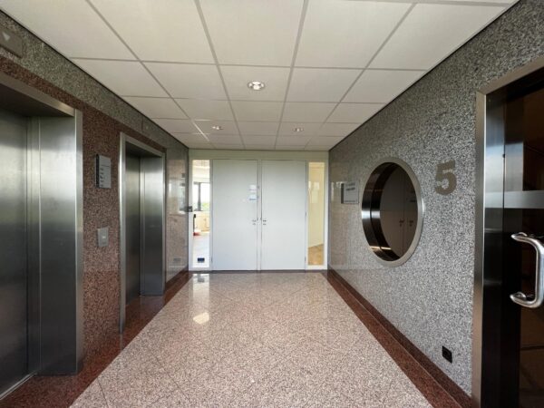 Hal met twee liften, een witte deur en het cijfer 5 op de muur in een kantoorgebouw aan de Nevelgaarde.