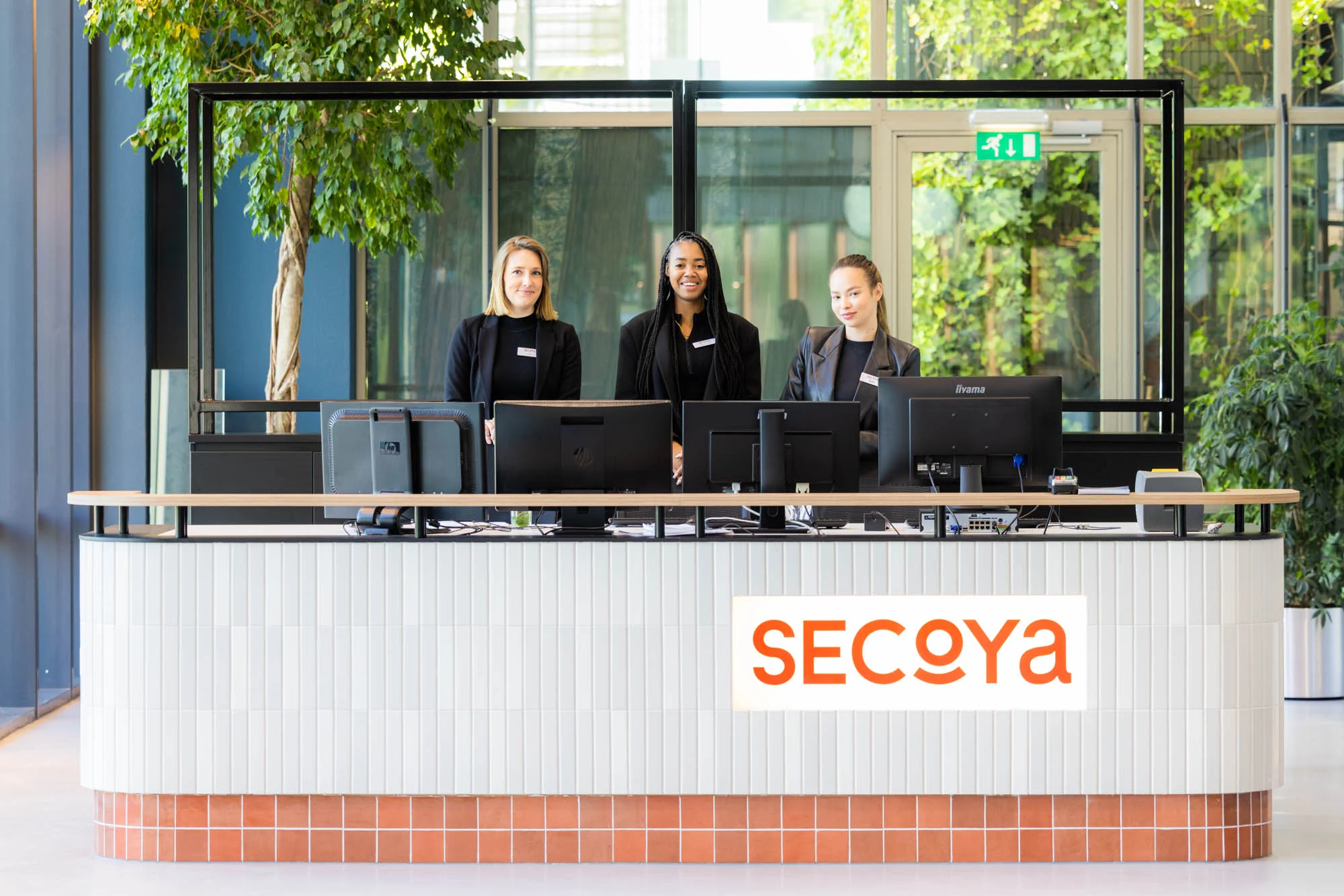 Drie receptionistes staan achter een moderne balie met het logo van Secoya in een lichte, groene ontvangsthal aan de Papendorpseweg.