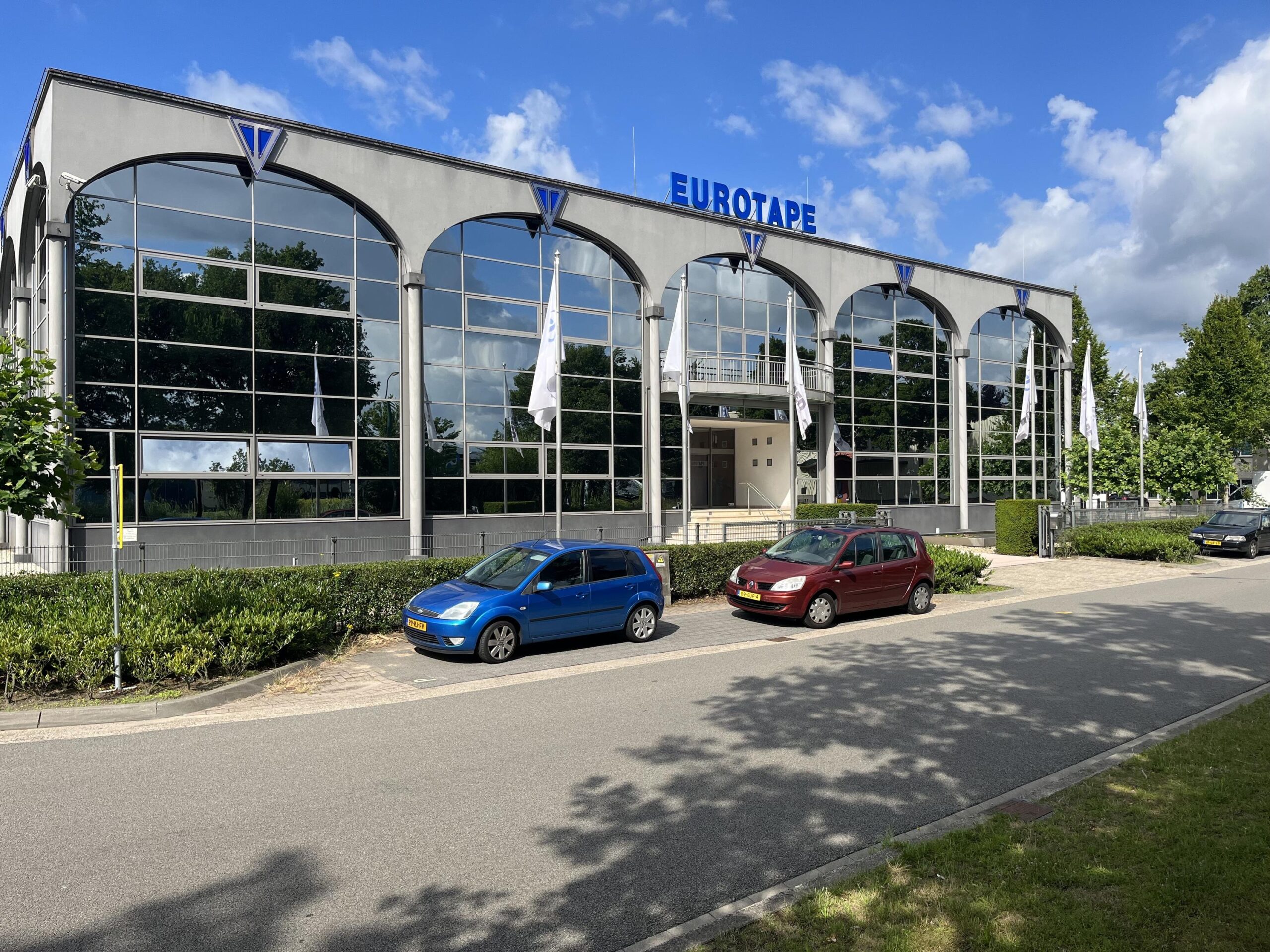 Voorzijde van een modern kantoorpand met de naam "EUROTAPE" erop en geparkeerde auto's op de oprit.
