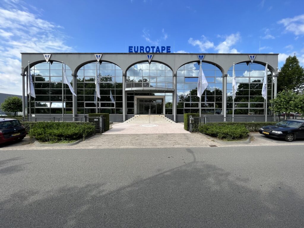 Het beeld toont de voorgevel van het Eurotape-gebouw met glazen panelen, vlaggen en een parkeerplaats ervoor.