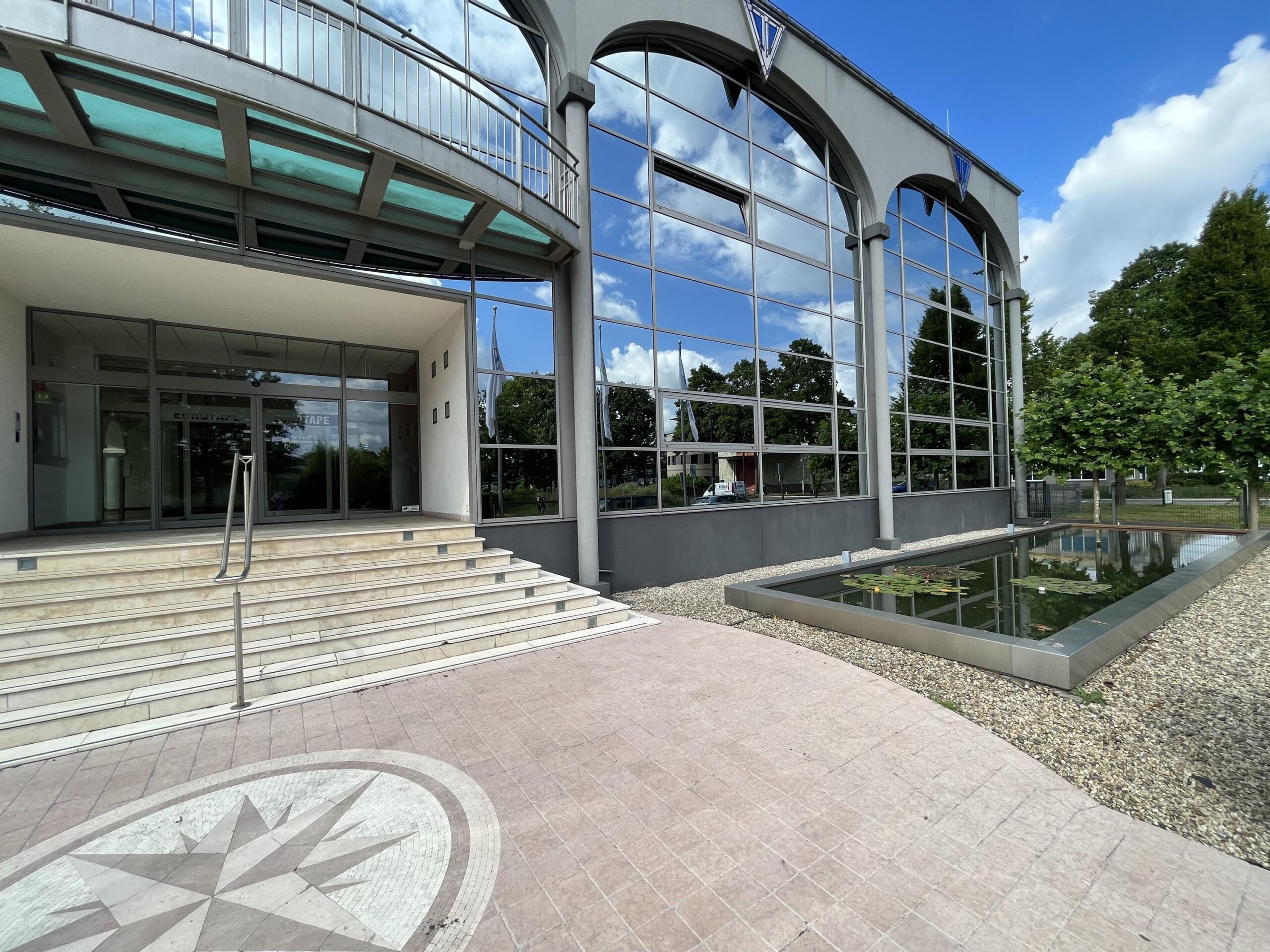 Ingang van een modern gebouw met glazen gevel en een vijver ervoor onder een blauwe lucht.