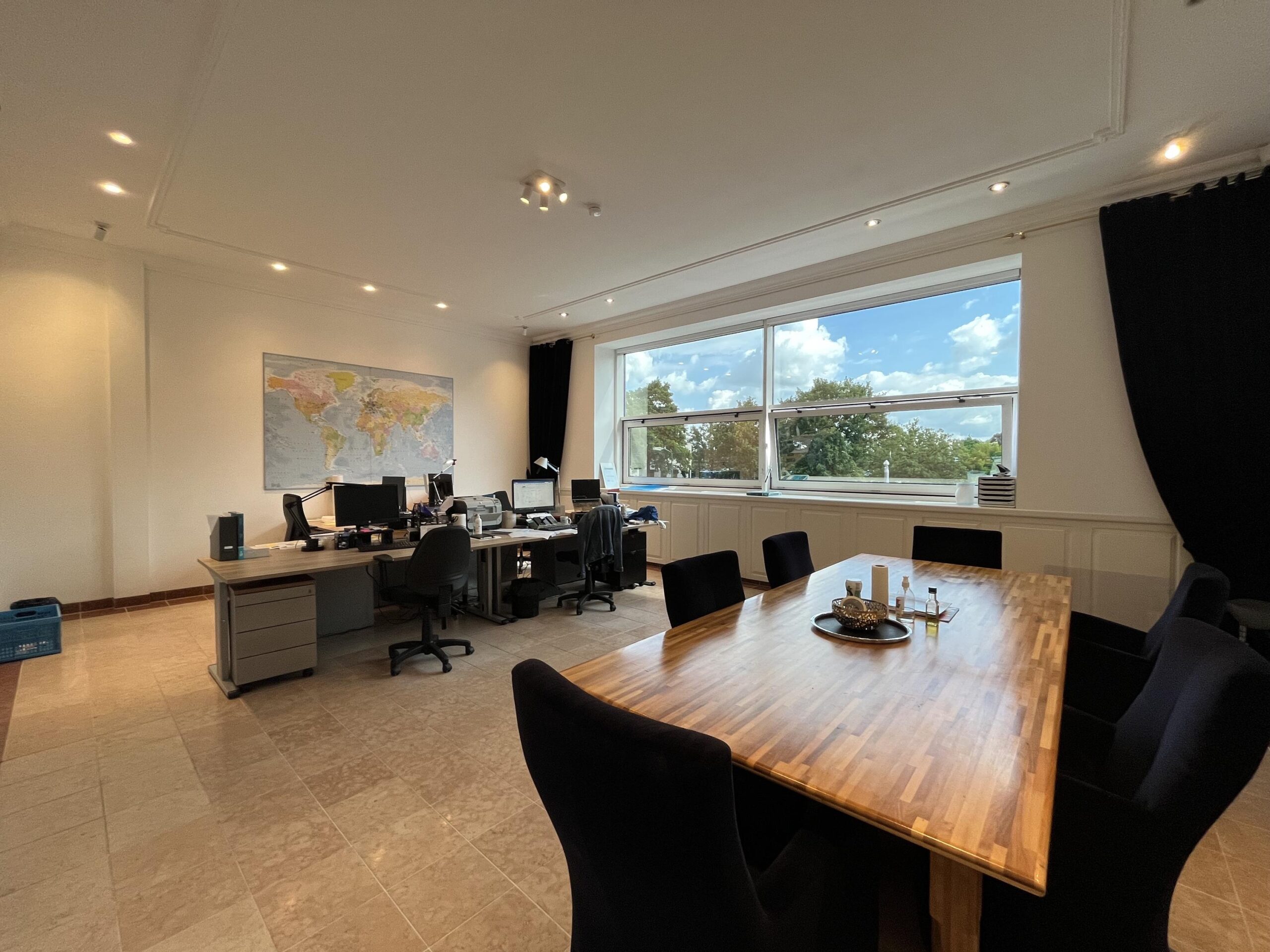 Een moderne kantoorruimte met een grote vergadertafel en bureaus met computers, verlicht door natuurlijk licht van een groot raam met uitzicht op bomen.