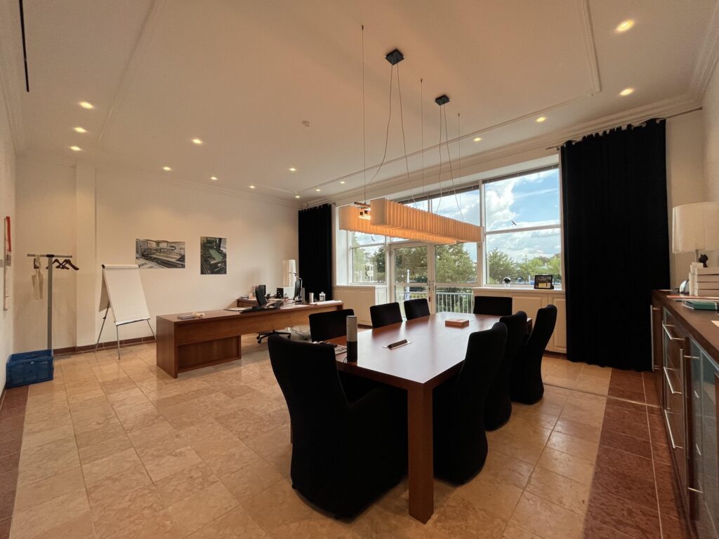 Een moderne vergaderruimte met een grote tafel, zwarte stoelen, en grote ramen met uitzicht op bomen.